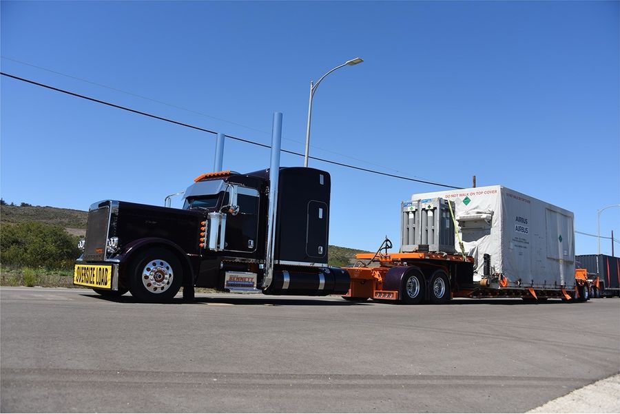 A long, dark purple semi-truck with chrome accents and a yellow "OVERSIZE LOAD" banner on the front bumper is parked on a paved road. The truck is hauling a large piece of cargo on a trailer, which is covered by a white tarp with the "AIRBUS" logo. Several tall, light-green gas cylinders are strapped to the front of the cargo. The scene takes place on a clear, sunny day with rolling, grassy hills in the background.