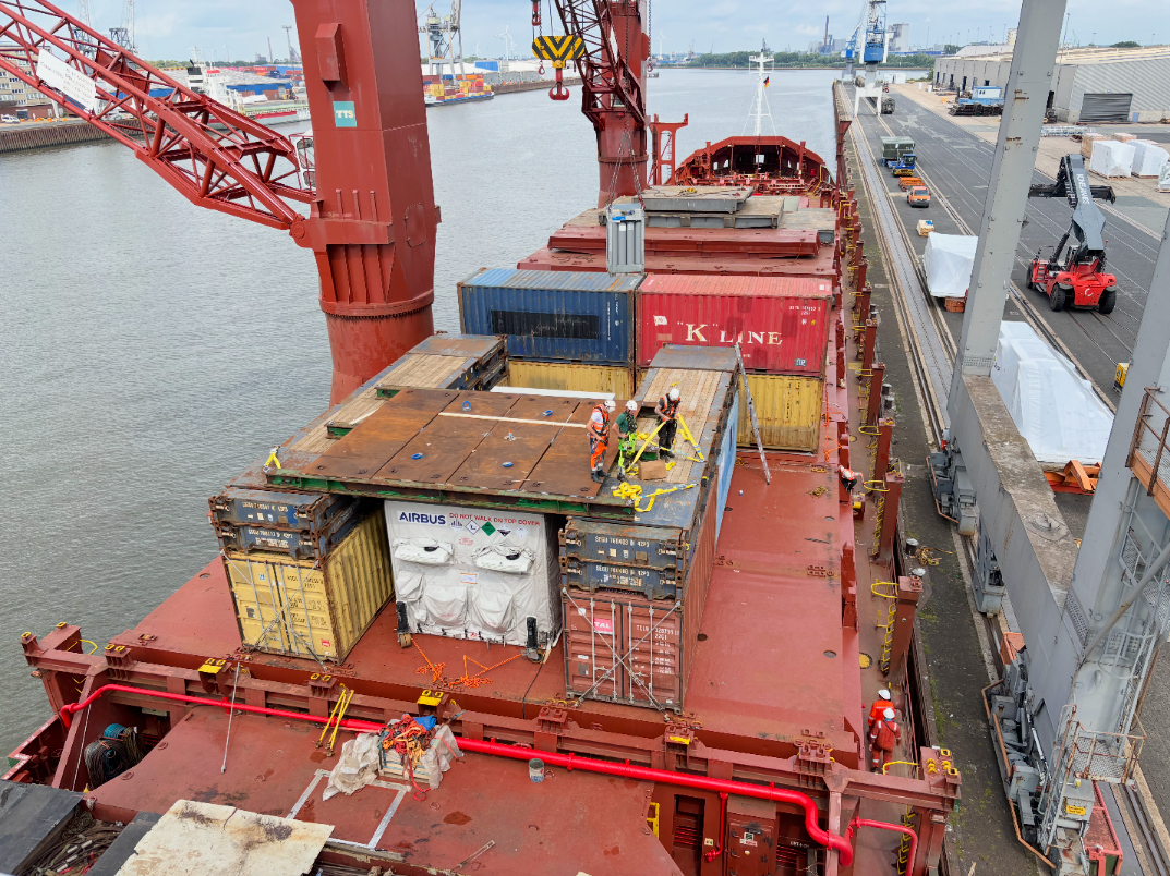 An overhead, wide shot of a large, red cargo ship docked in an industrial port. On the deck, several workers in hard hats and safety gear are securing a large, white-wrapped cargo container with the Airbus logo. The container is positioned on top of and between other shipping containers. The workers are using yellow straps to fasten it down. The background shows the busy port with water, cranes, and other docked ships.