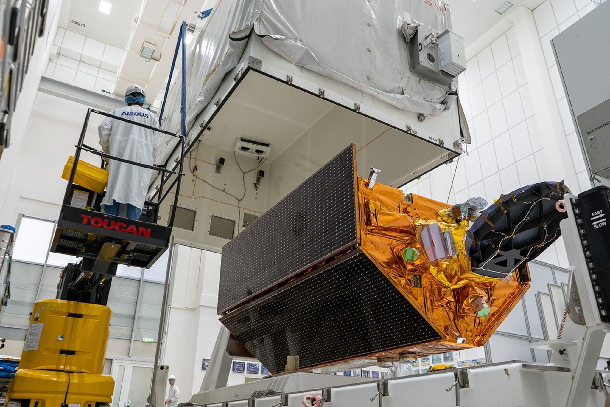 A low-angle shot from inside a large, white cleanroom shows a satellite being enclosed in its shipping container. The satellite, covered in gold-colored thermal blankets and with large, dark solar panels folded against its sides, is suspended in the middle of the room. A large, white upper section of a container is being lowered down over the satellite from above. To the left, a technician dressed in a full white cleanroom suit with the "Airbus" logo stands on a yellow aerial work platform, monitoring the procedure.