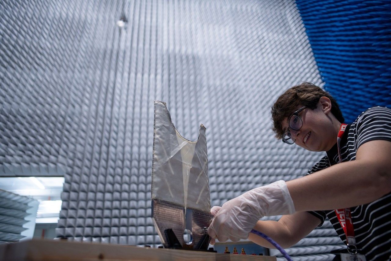 A gloved person in striped shirt and glasses attaches a blue cable to a device with a metallic base and a translucent-appearing pane. The background is a chamber lined with grey and blue geometric foam acoustic panels.