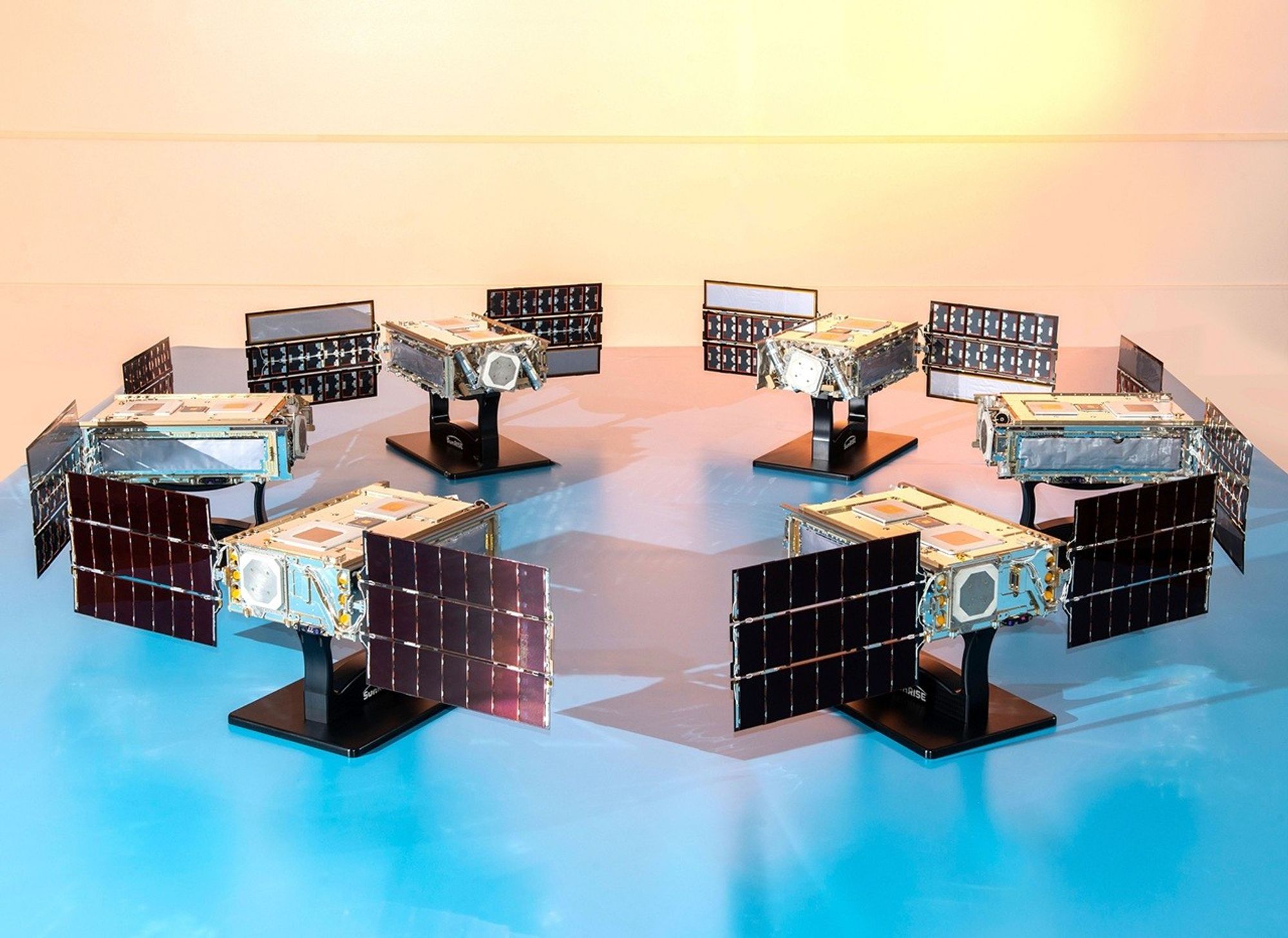 Six identical CubeSats arranged in a semi-circle on a reflective blue surface, with their solar panels deployed. Each small, boxy satellite is mounted on a black stand. The background is a smooth, light-colored wall, illuminated with a warm, bright light from the upper right, mimicking a sunrise.