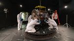 People in cleans suits work on a spacecraft in a large chamber.