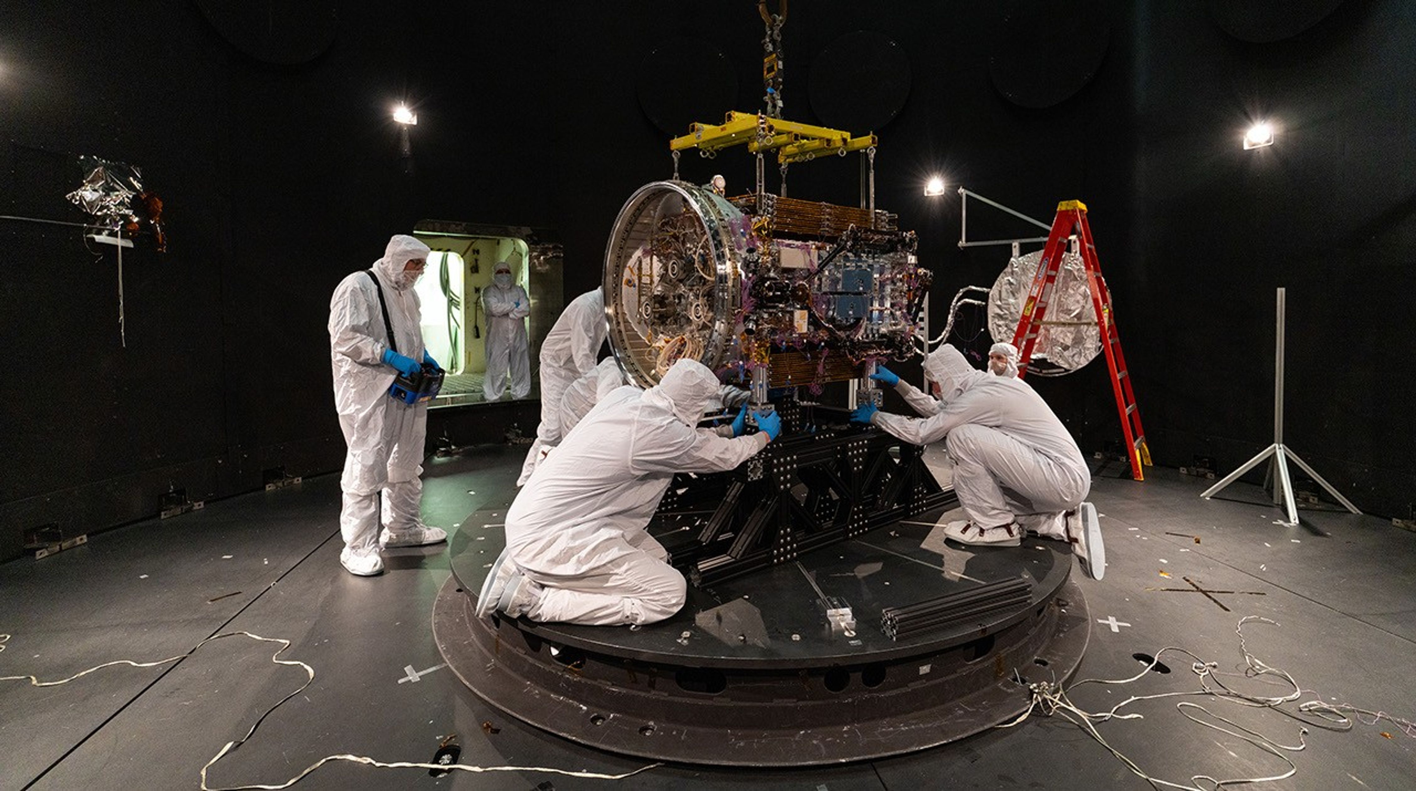 People in cleans suits work on a spacecraft in a large chamber.