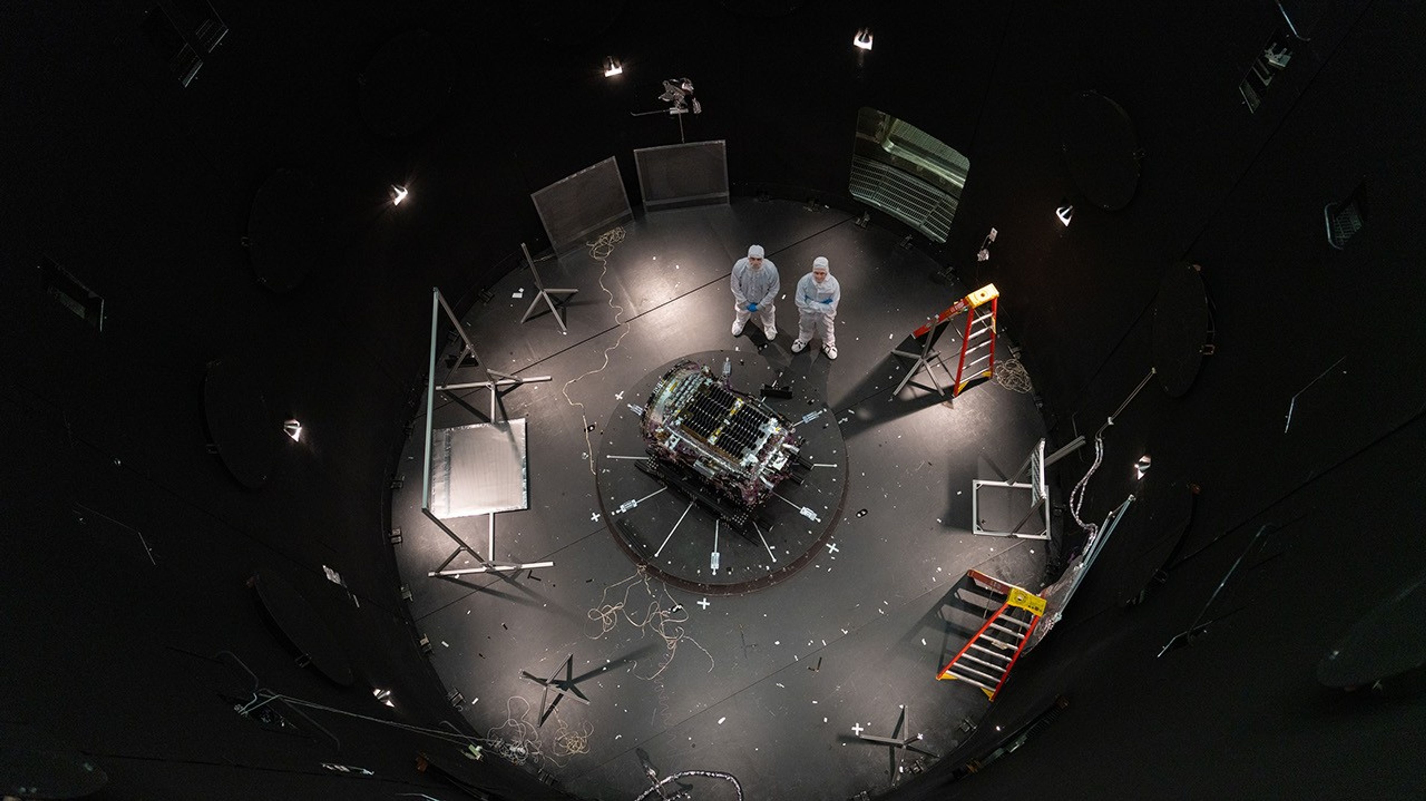 Two people stand next to a spacecraft inside a large black chamber.