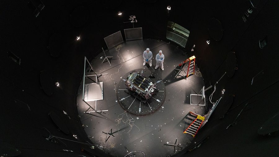 Two people stand next to a spacecraft inside a large black chamber.