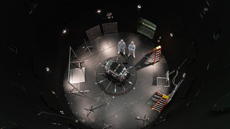 Two people stand next to a spacecraft inside a large black chamber.