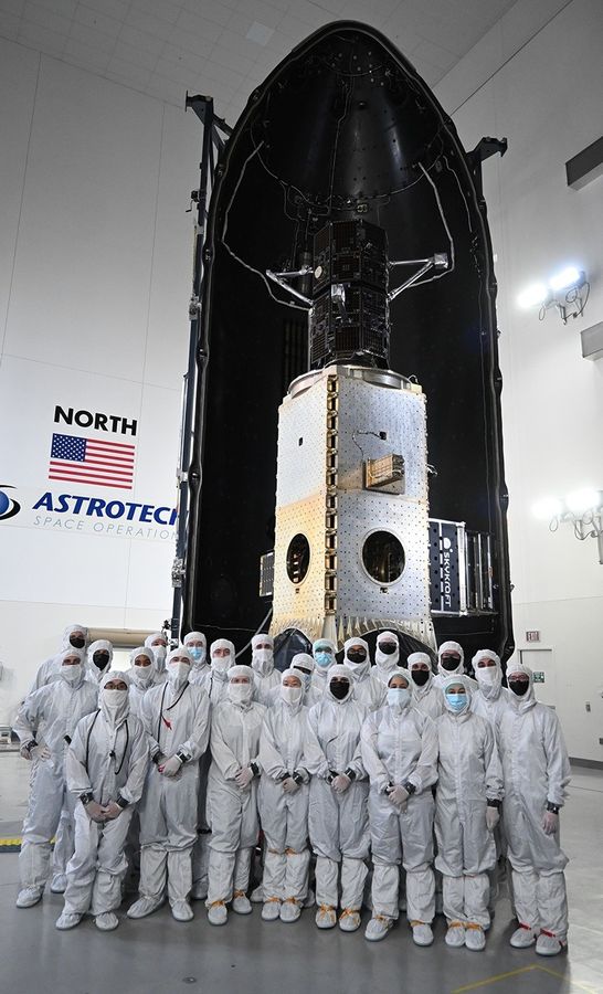 Image shows a group of people in white bunny suits posing in front of a spacecraft fairing with two black satellites on top a shiny metallic box