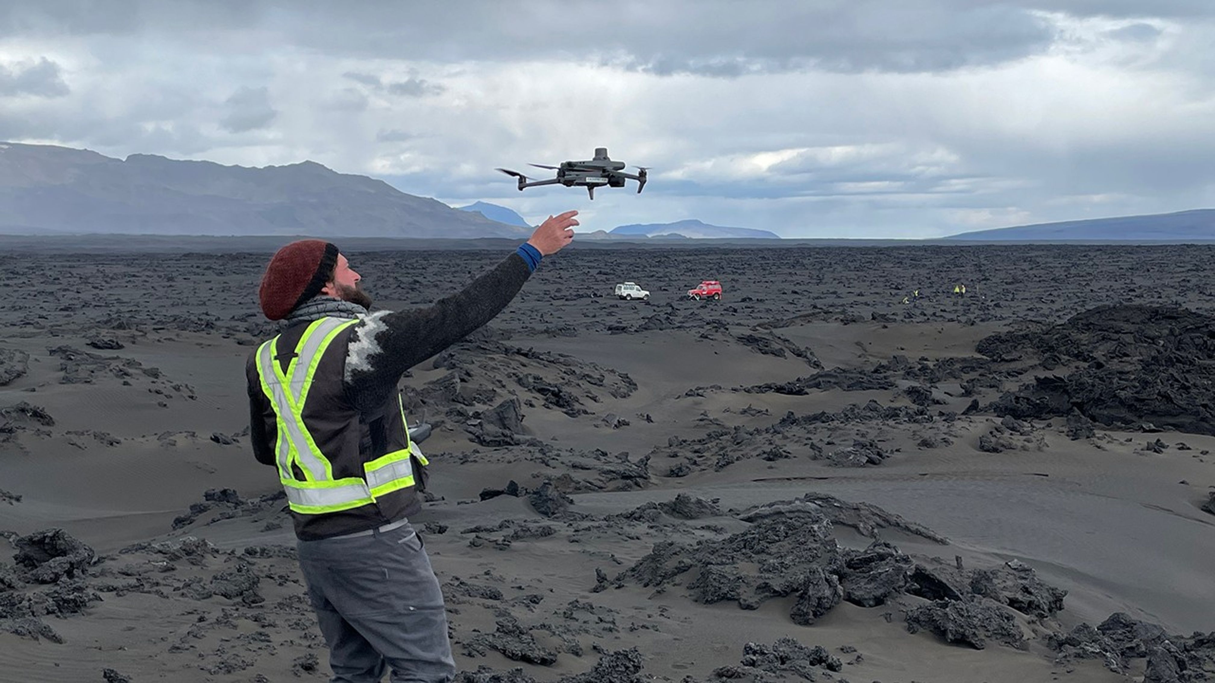 A man with a red cap and a bright green reflective vest holds his right arm out above a barren region in Iceland. A drone is just above his arm. The sky is partly cloudy. There are mountains in the distance.