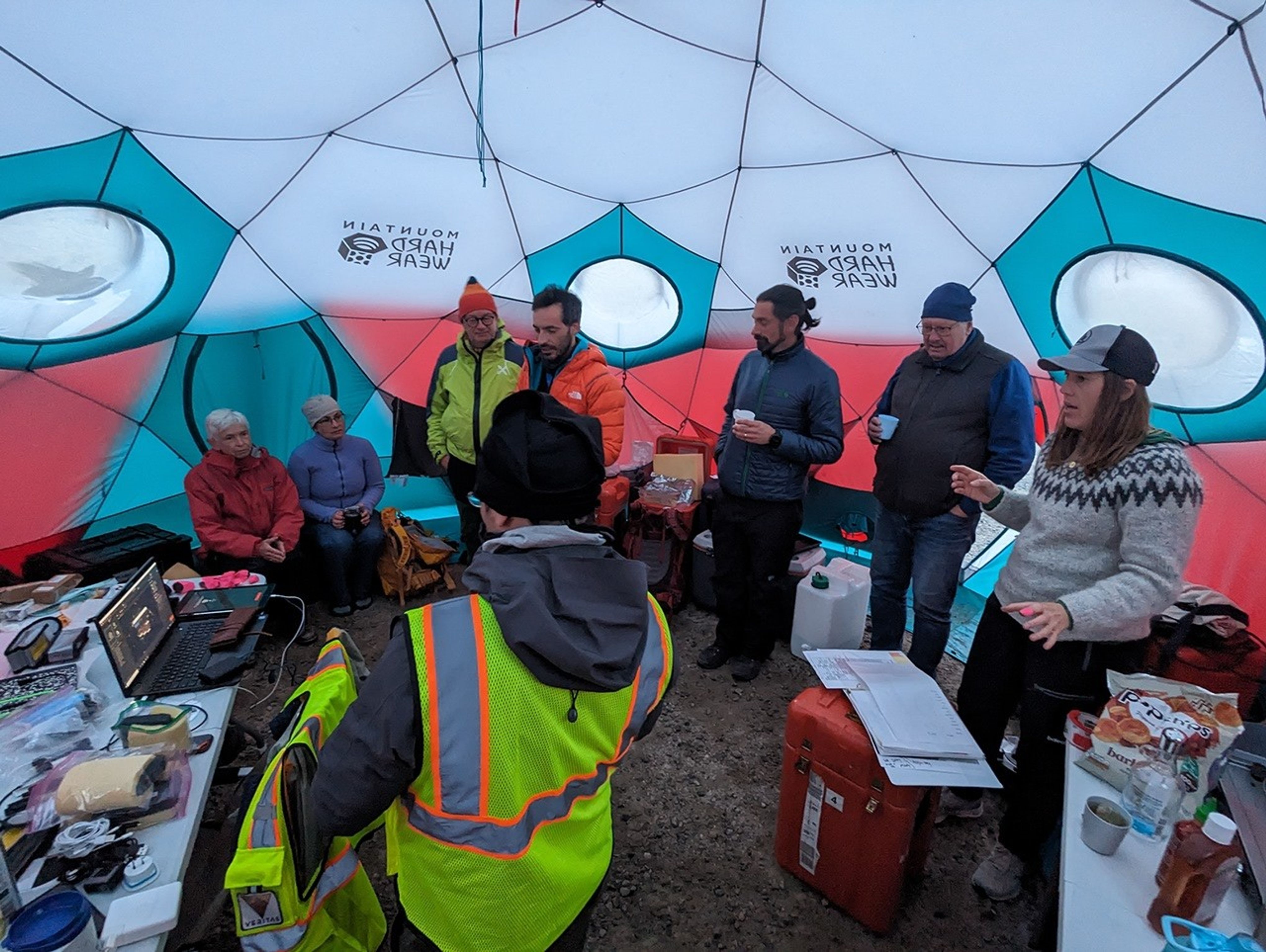 People with cold weather clothing stand and sit in a tent. Some of the people are holding mugs or cups. A table with laptops and gear is off to the left of the image. A table with food is to the right.