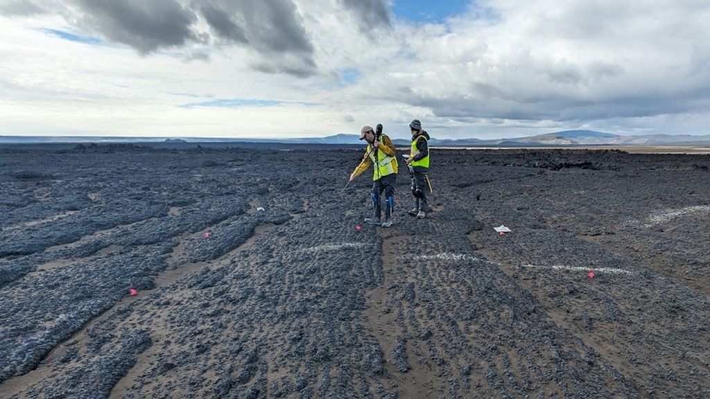 Two people wearing bright green reflective vests stand in a rocky field .