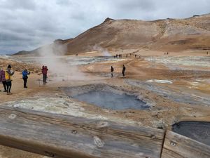 Several people take photos and walk around craters from volcanoes. The land is reddish and gray with what looks like ponds with steam rising out of them.