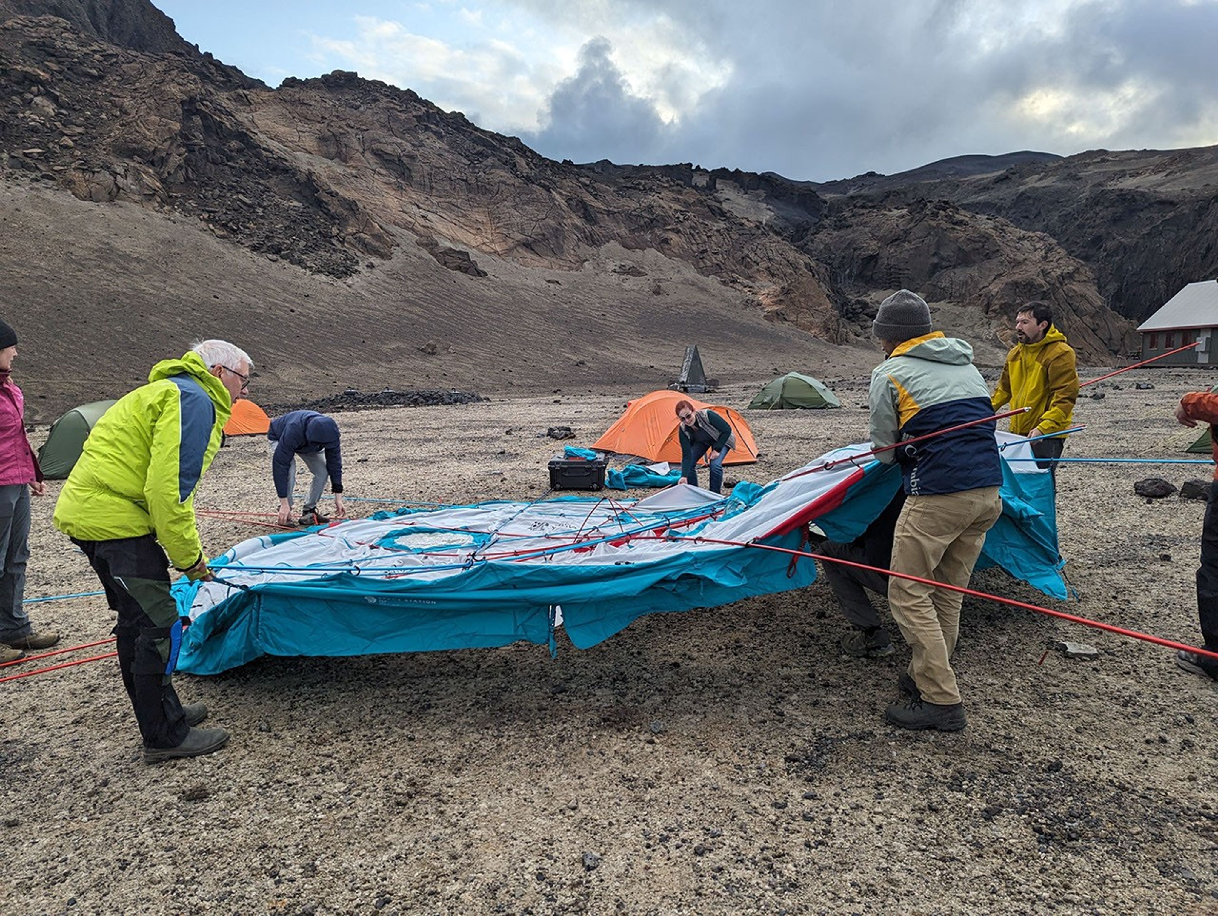 People wearing cold weather gear hold onto a round blue tent with tiedown ropes attached.