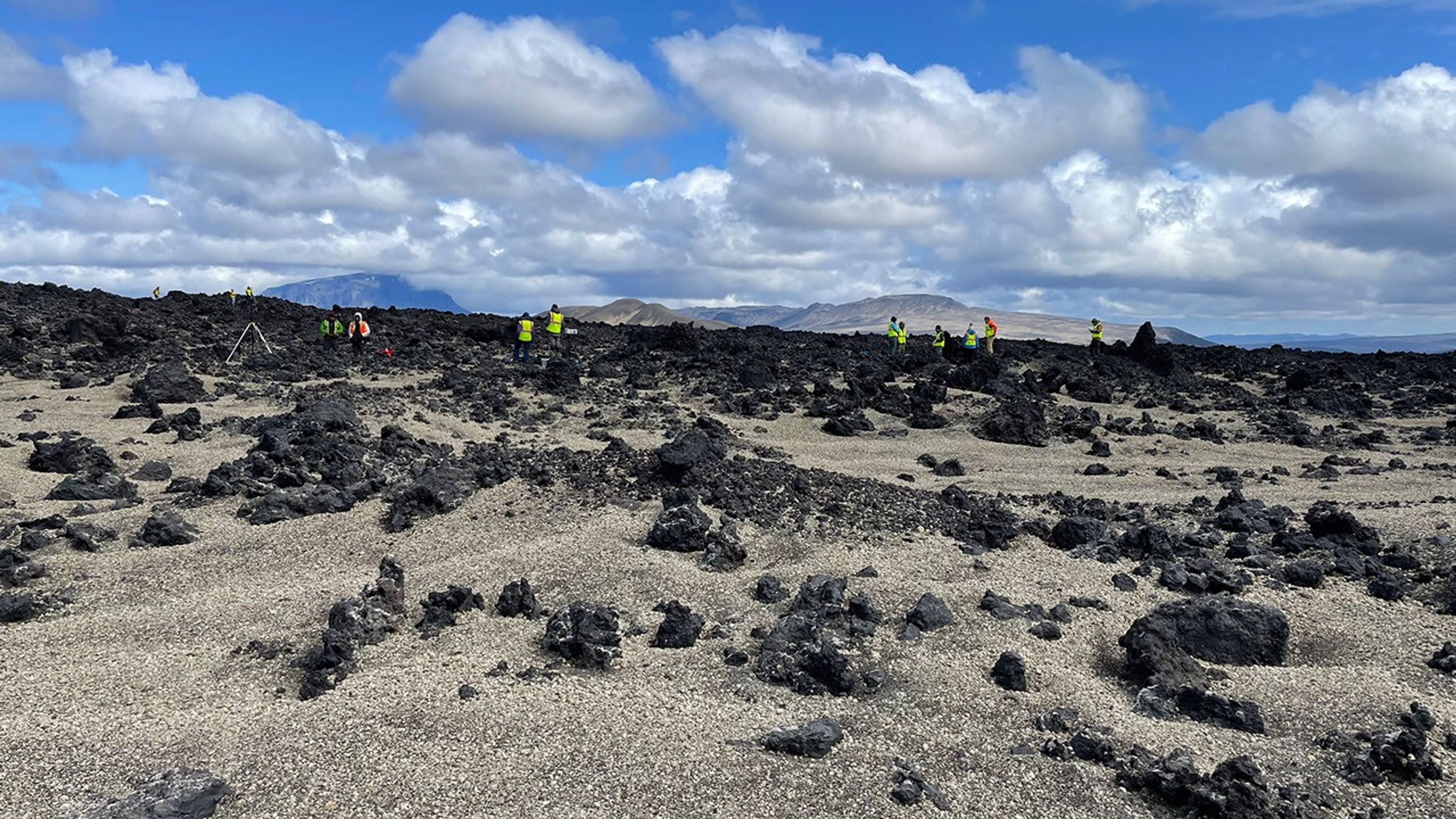 People in bright green vests can be seen in the distance with a mountain in the distance. The ground is rocky with scattered green vegetation.