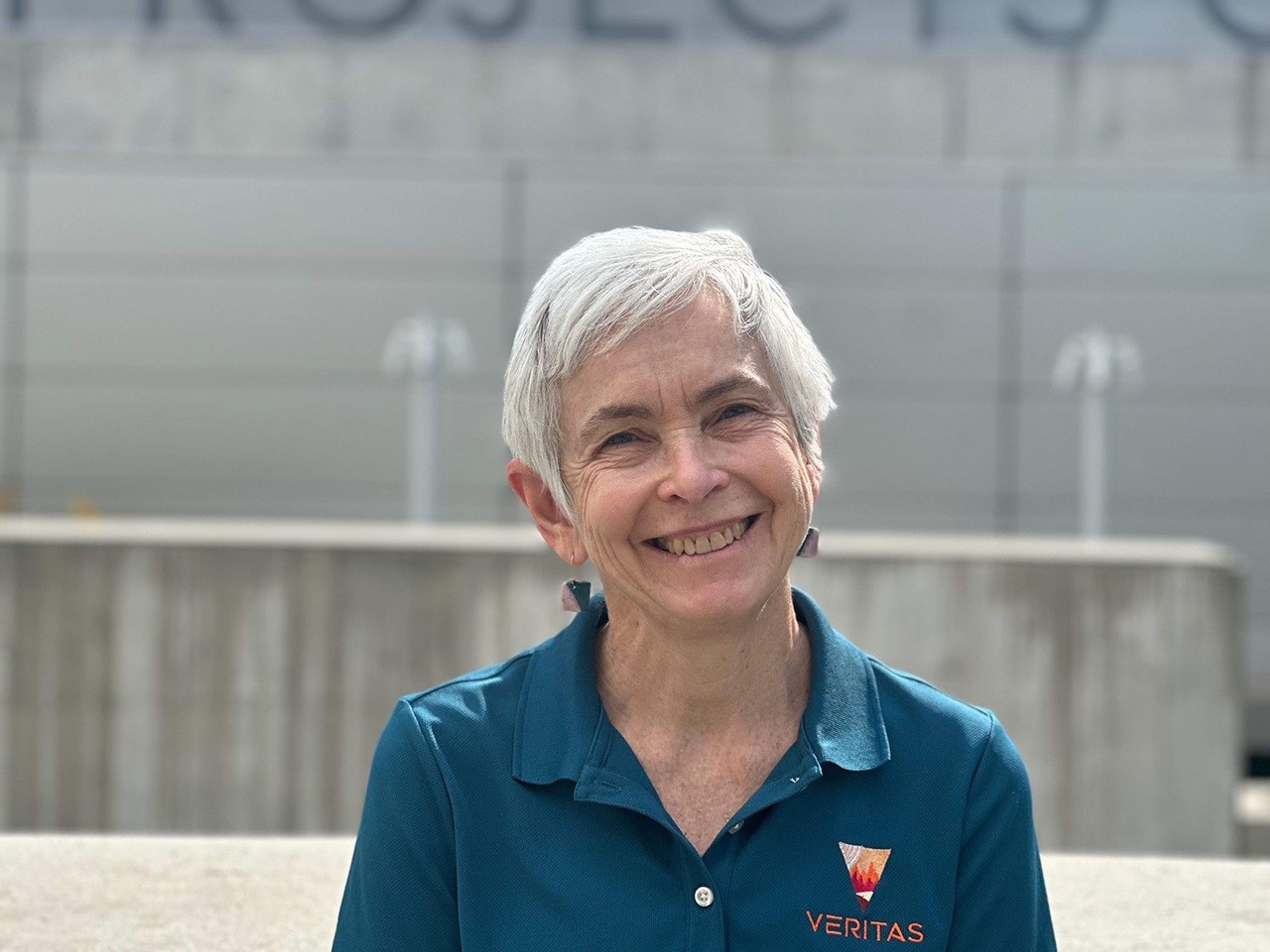 A color portrait photo of a smiling woman looking at the camera, with he head tilted slightly to the left, wearing a teal polo shirt with a logo embroidered on her left chest, an orange and red upside-down triangle with the word VERITAS below. Behind her is an out-of-focus gray concrete building filling the frame.