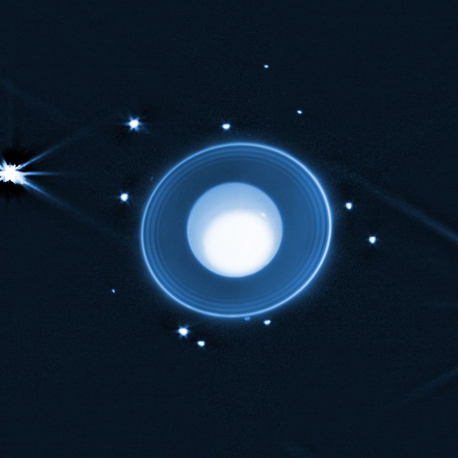 At center of this is the planet Uranus, a blue globe with a large white spot occupying most of the planet. It’s surrounded by a series of rings that get brighter as you move outward. Beyond the rings are 14 moons appearing as points of light. Just beyond the outermost ring at 10 o’clock, a faint moon is visible.
