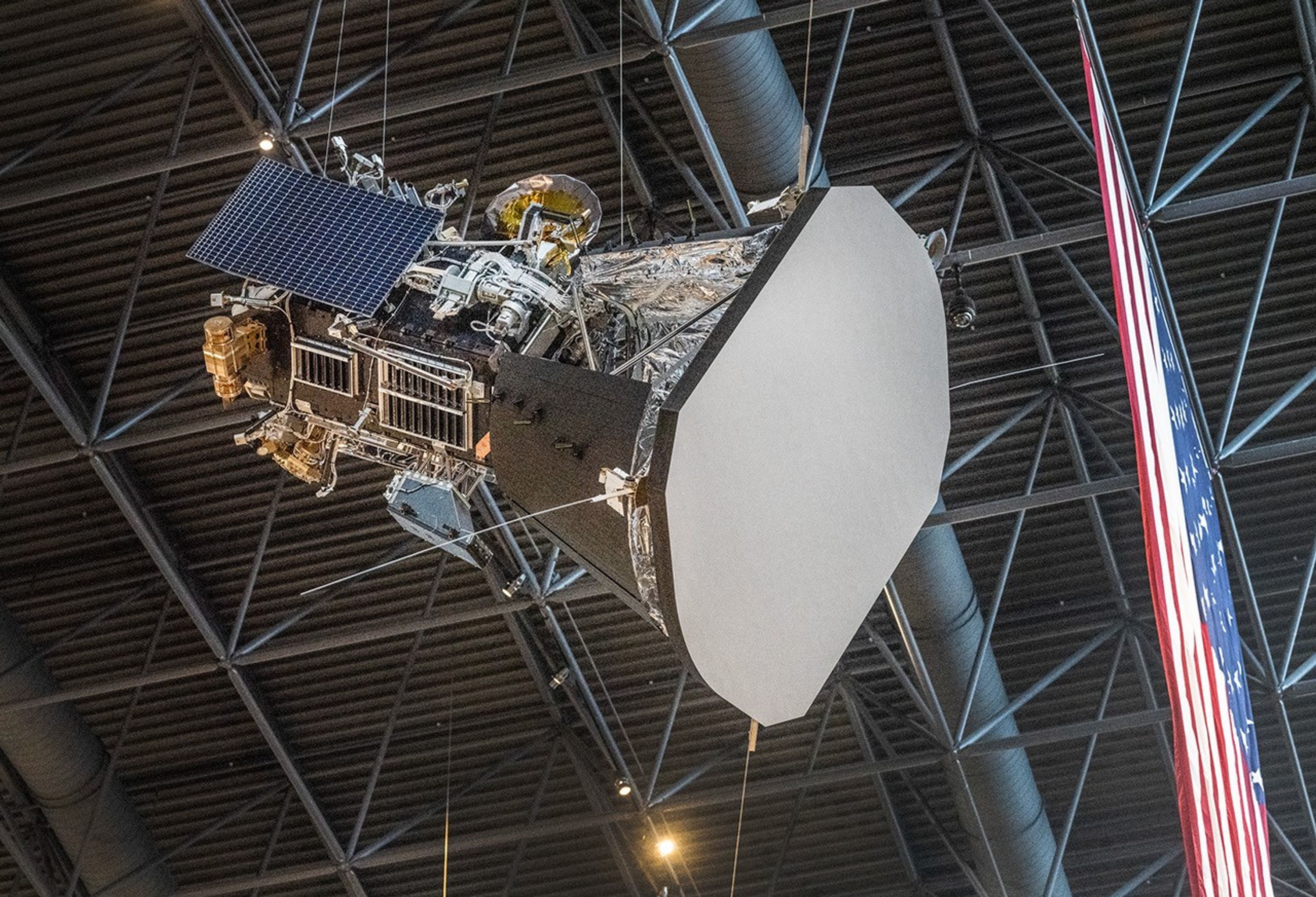 The Parker Solar Probe model is suspended from the ceiling by cables. The side view of the spacecraft shows one of its solar panels, the heat shield, and several other instruments and systems. Part of an American flag is visible hanging next to the model.