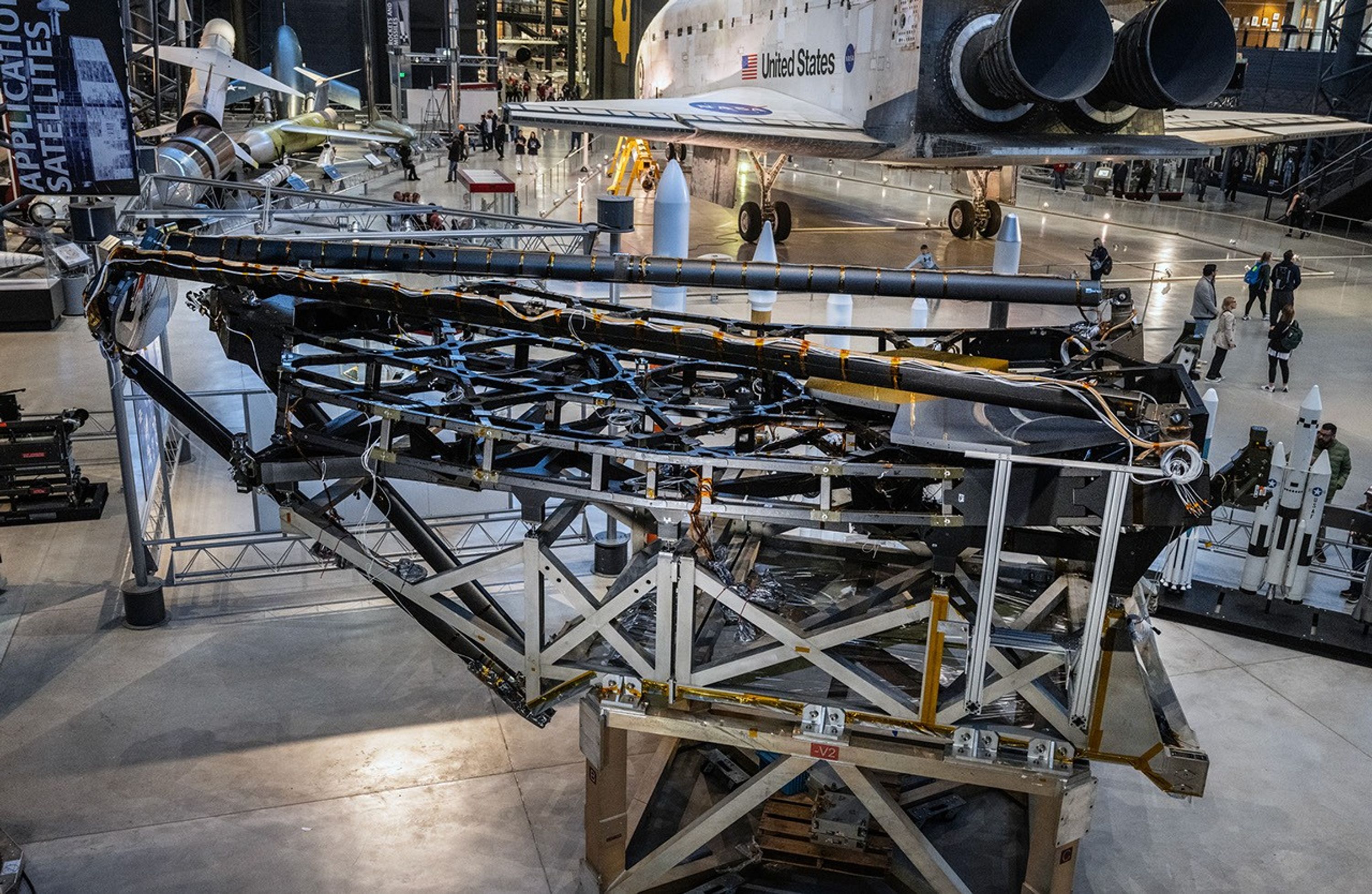 Located in a large open room with grey cement floors, backdropped by a large white and black Space Shuttle with “United States” and an American flag on its side. The James Webb Space Telescope Optical Telescope Element Pathfinder sits on top of a large silver stand with cross braced supports. The pathfinder is made of black carbon fiber material with gold and silver wiring wrapping all around the structures dark black and grey components. On the lower right side of the pathfinder two large hexagonal mirrors rest on top of the black hexagonal support structure below it. One mirror shines in gold, and the other in silver. Above the mirrors two long black support arms reach upward over the left-hand side of the pathfinder, connecting to a third support arm that all are holding a circular silver mirror that is designed to fold out in front above the other two mirrors. Behind the pathfinder a series of different sized and configured rocket models.