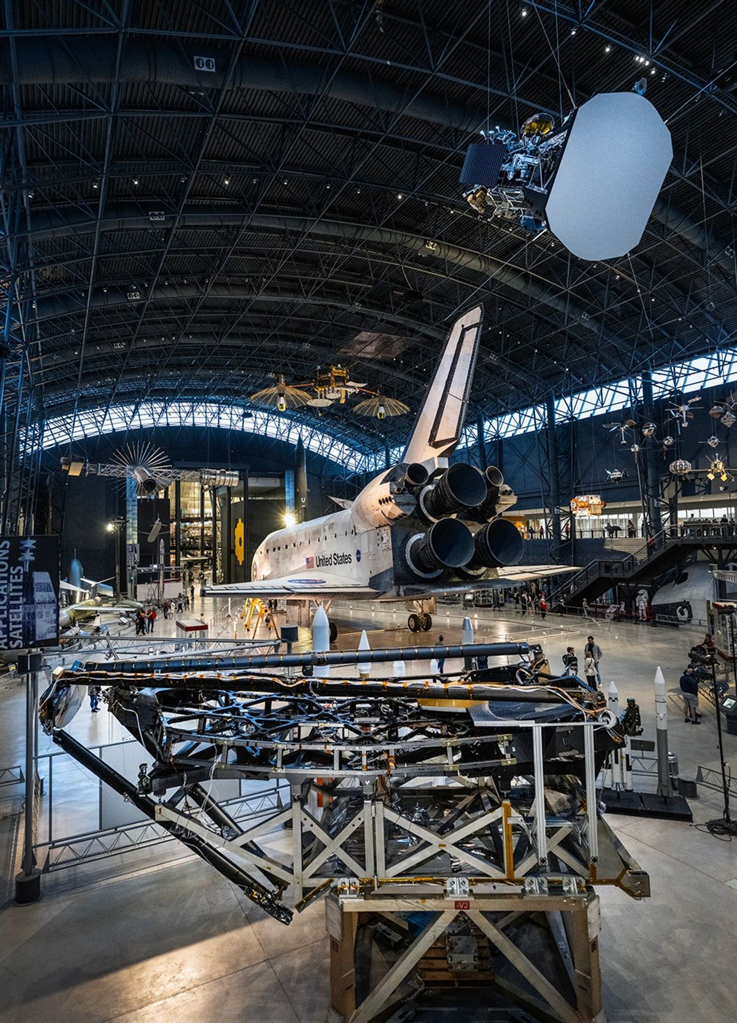 Located in a dark grey, high ceiling airplane-like hangar. A line of bright windows lines the upper part of the hangar, just below the ceiling. In the center of the image a large white and black Space Shuttle takes up most of the image, with three large conical engines in a triangular pattern on its back, with “United States” and a large American flag on its side. Hanging from the ceiling in front of the Space Shuttle is a model of the spacecraft Parker Solar Probe which has a large grey shield at its front that is curved at two ends and flat on the other two, with antenna pointing out from each of the four ends of the shield. Behind the shield a mix of shining metallic materials, and a blue square solar array with silver lines running through it. On the floor in the bottom left corner, a large metallic framework nearly 30 feet wide holds a black carbon fiber structure above it with a series of hexagonal frames on the structure. Above two of the hexagonal frames, two large hexagonal mirrors face upward, one shining in gold, and the other in silver. Above the hexagonal frame and mirrors, three large arms made of the same black carbon material rest over the top, holding a different silver circular mirror that is designed to fold out in front of the hexagonal frame, but is currently stowed. The large open room hangar has many additional smaller bright silver and gold satellites of varying size also hanging from the ceiling in the background.