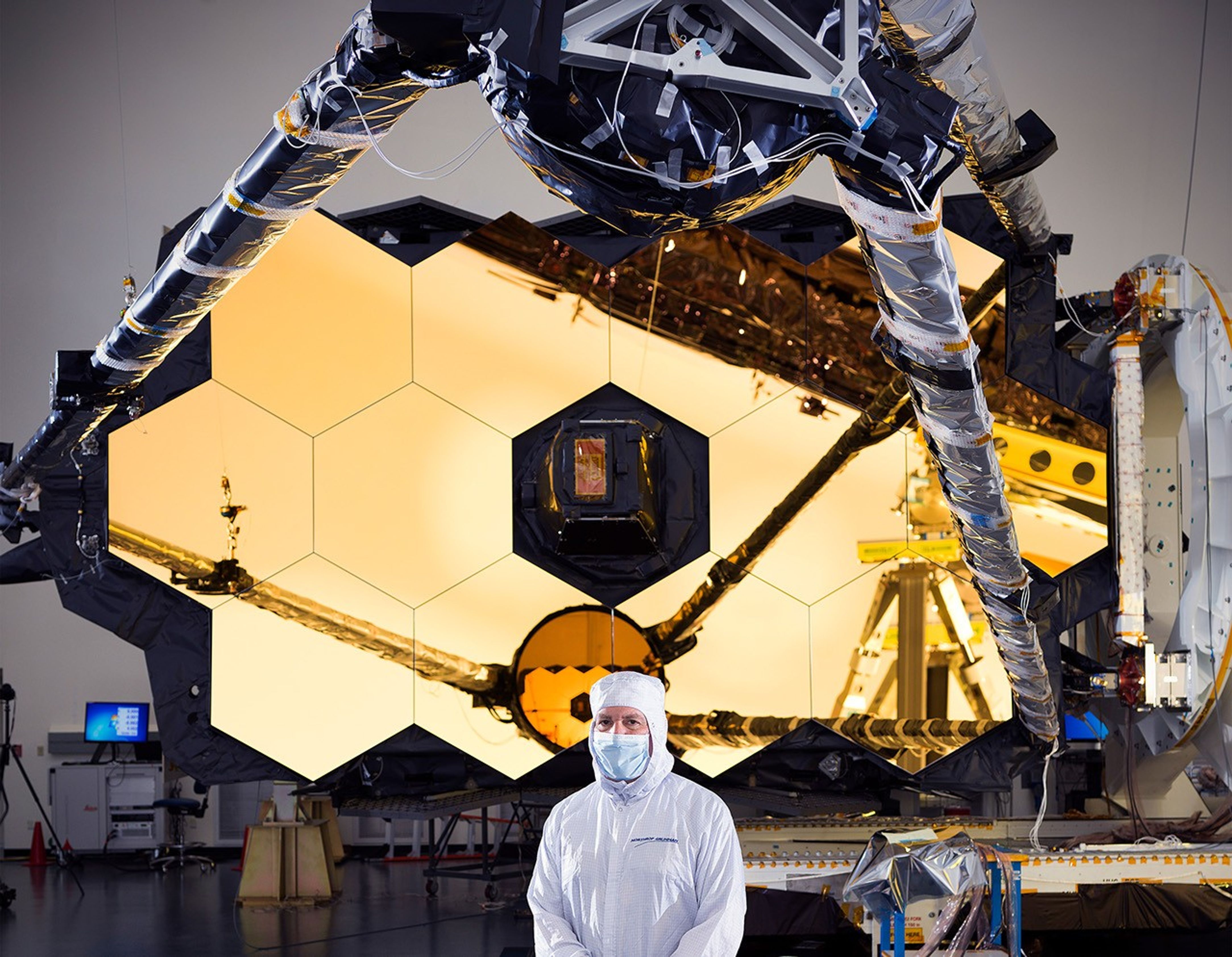 Lee Feinberg stands in front of Webb Space Telescope Optical Telescope Element (OTE) comprised of the haxagonal primary mirror segements with the round secondary mirror reflected in the primary mirror.