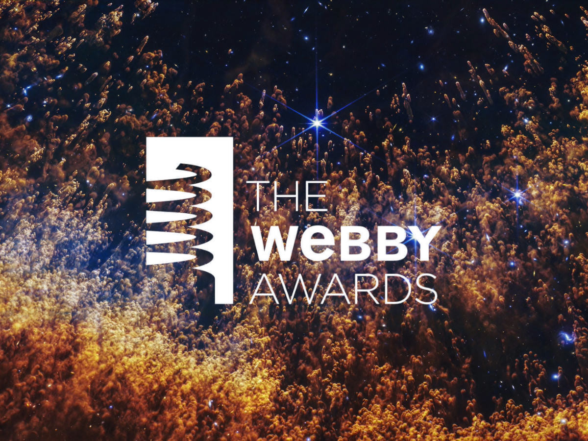 A closeup of a small section of the Helix Nebula, an expanding shell of gas and dust. Thousands of orange and gold comet-like pillars stream upward from the bottom, like thin liquid blown up a sheet of glass. These pillars are around the circumference of the arced shell, which forms a partial orange semi-circle at the bottom. The pillars are more numerous and denser at the bottom, and darker red. They fade to orange and then yellow in the arc. In the top two-thirds, they are thinner and more golden, and it’s easier to see the black background of space. Several bright blue stars, some with diffraction spikes, are scattered throughout. A few larger stars are on the right side. A white graphic with the words "The Webby Awards" and a graphic of the award is laid over the image in the center.