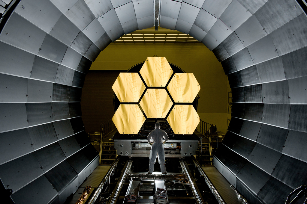 Fotografía de una persona de pie frente a seis segmentos de espejo hexagonales.