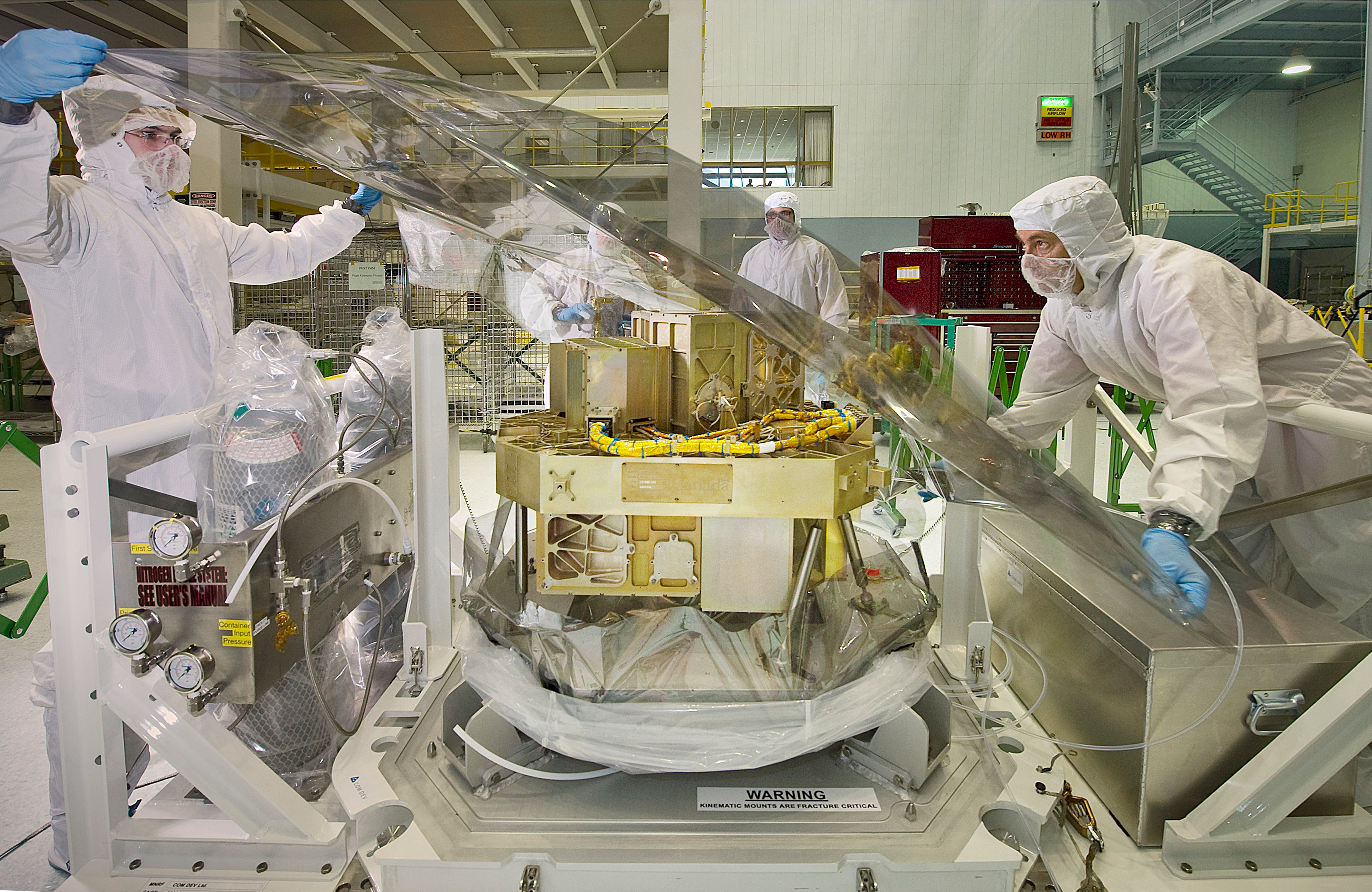 Photograph of Webb's Fine Guidance System (FGS) and Near-infrared Imager and Slitless Spectrograph (NIRISS) in a clean room. There are four people in white clean room suits with blue gloves.