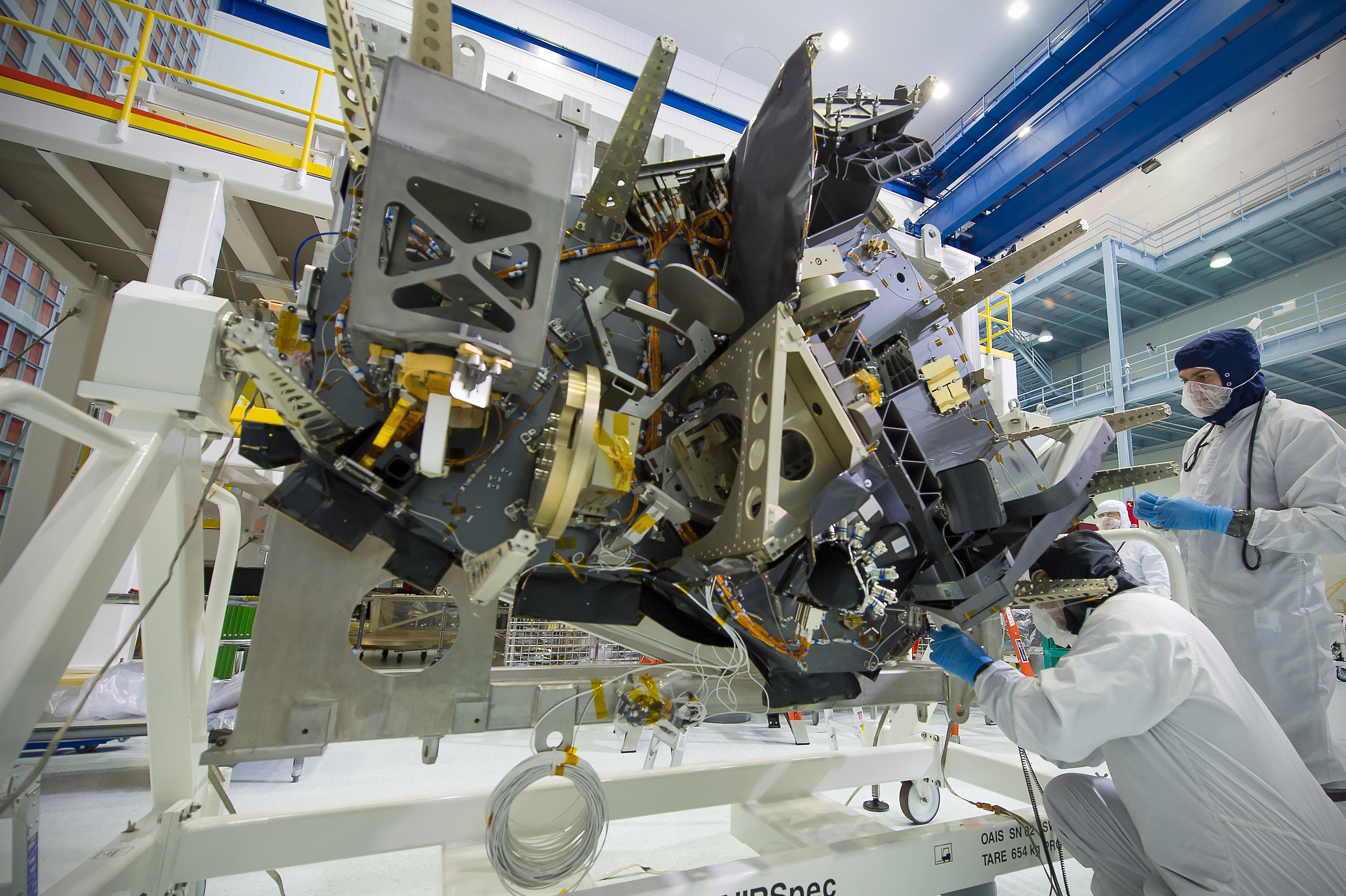 Photograph of Webb’s Near-Infrared Spectrograph (NIRSpec) instrument in a clean room. Two people in white clean room suits with blue gloves stand close to the instrument at right.