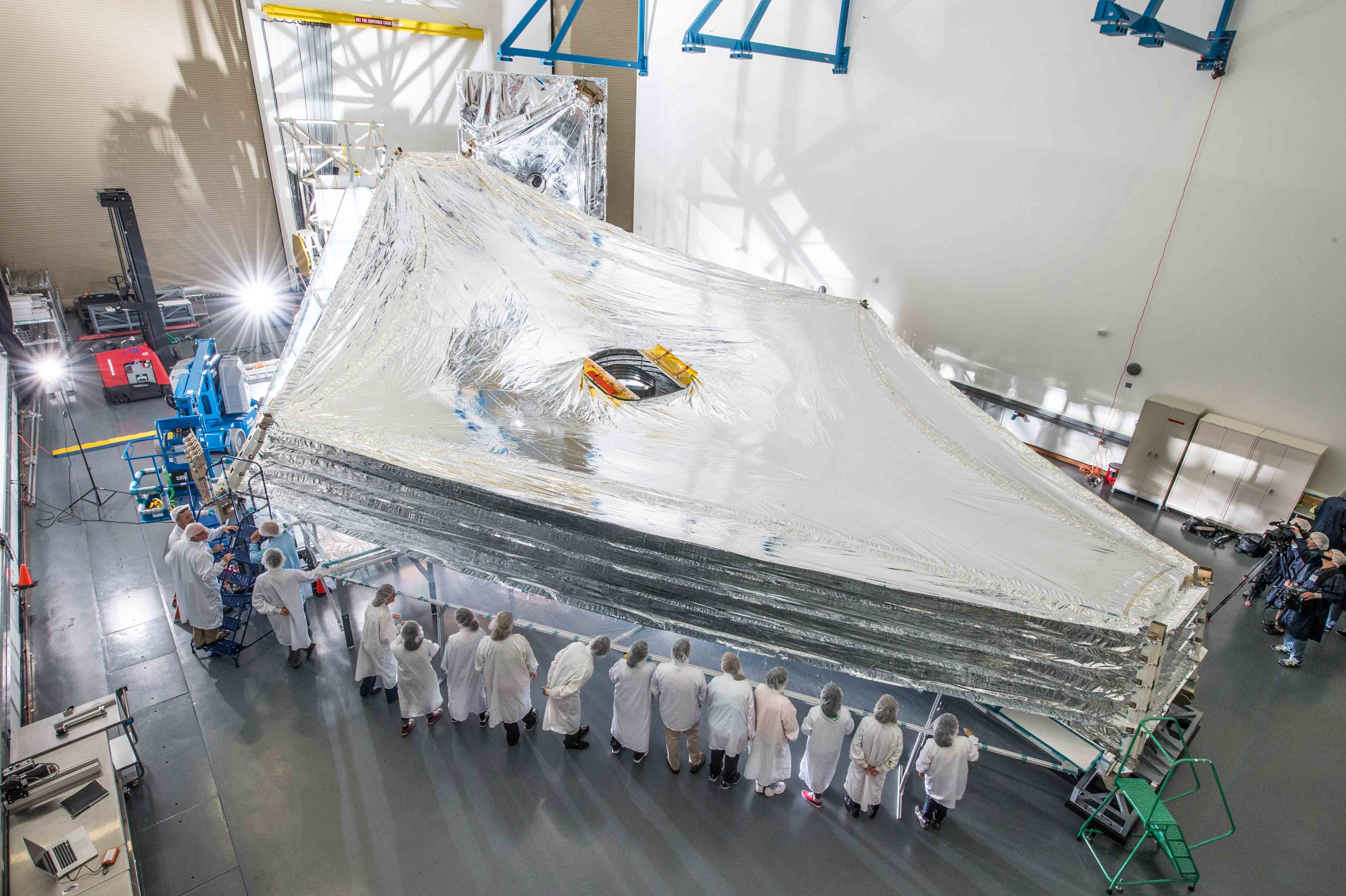 Photograph of the James Webb Space Telescope’s sunshield in a clean room. The sunshield is fully deployed, with spaces between each layer. White garbed people line the foreground side, and blue garbed photographers are in front of the sunshield.