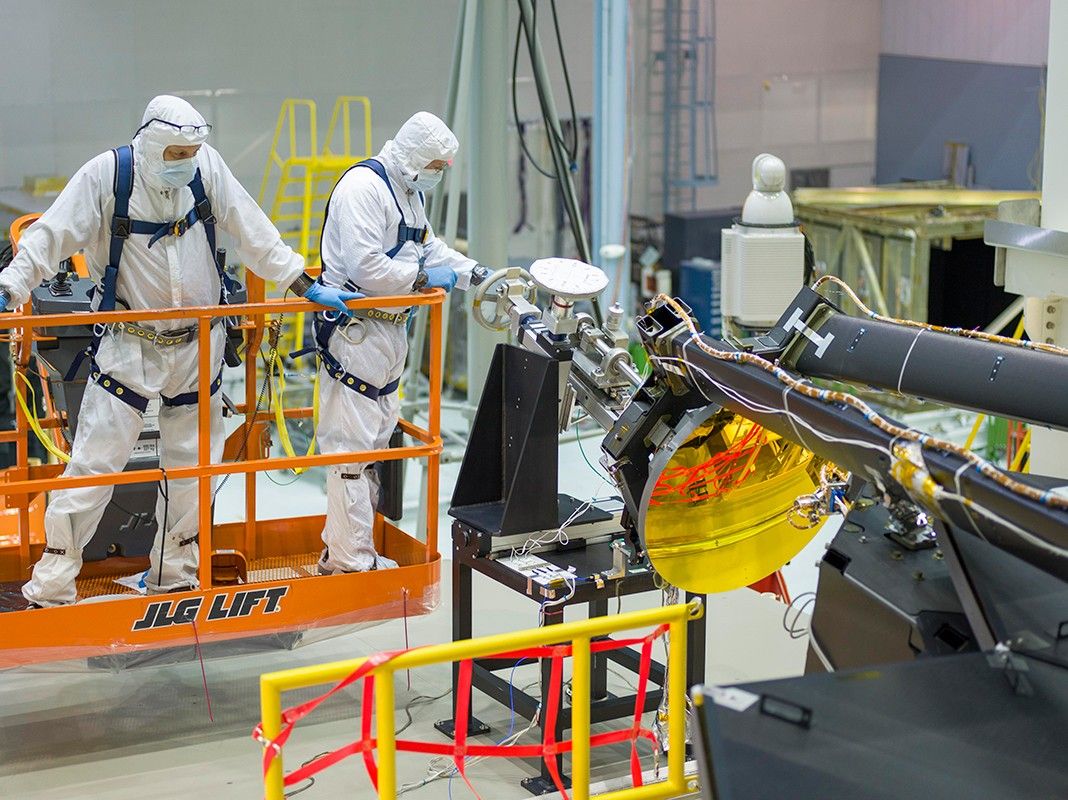 Photograph showing the installation of Webb’s secondary mirror in a clean room. Two people in white clean room suits with blue gloves stand on an orange lift at left.