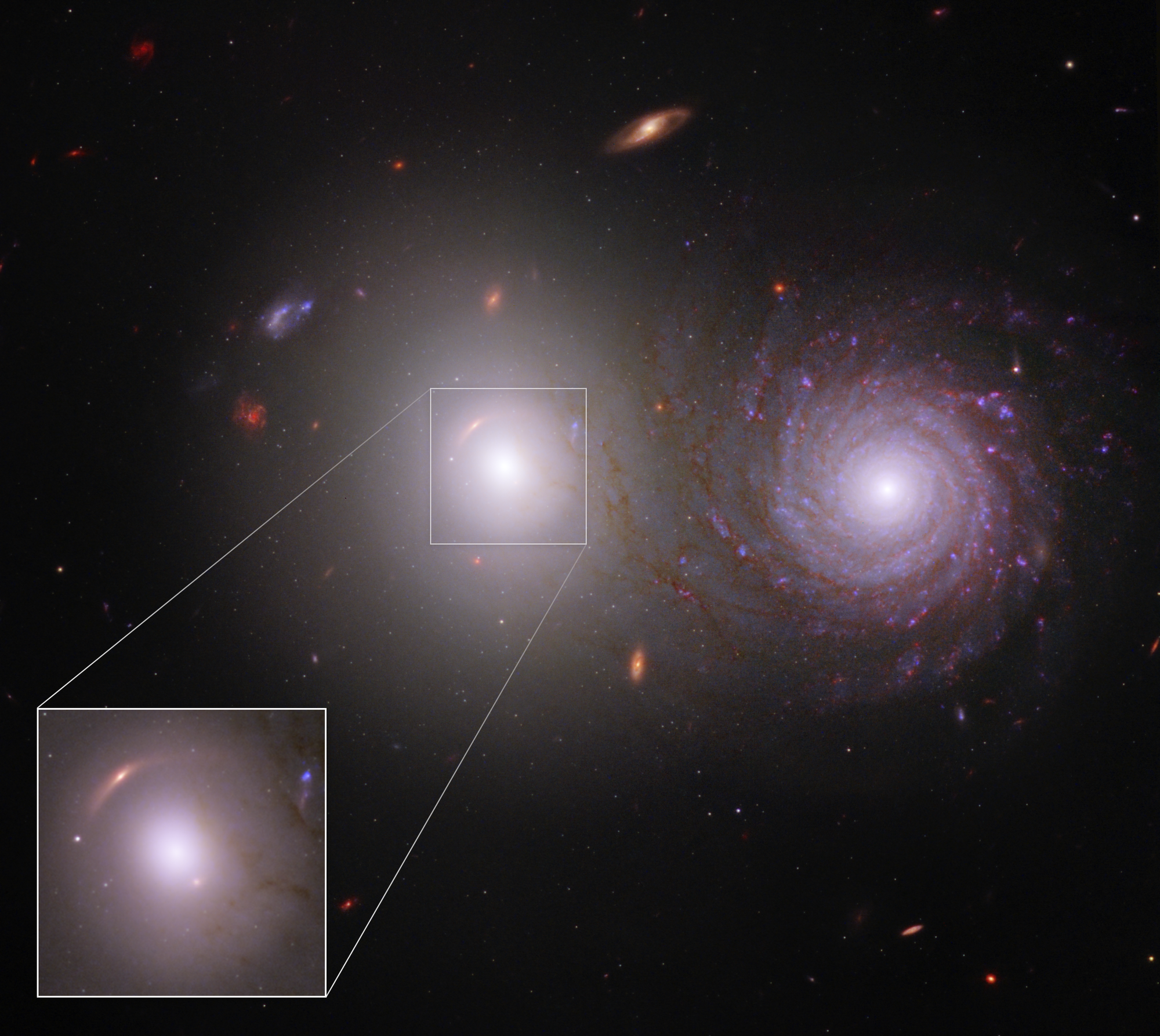 Two large, very bright galaxies dominate the center of the image. At right is a bright spiral galaxy. At left is an elliptical galaxy with an extremely bright core. A light white box overlays the elliptical galaxy and reappears at the bottom left, showing the area in a larger view. A stretched red arc appears above the elliptical galaxy at 10 o’clock and a red dot appears at 4 o’clock.