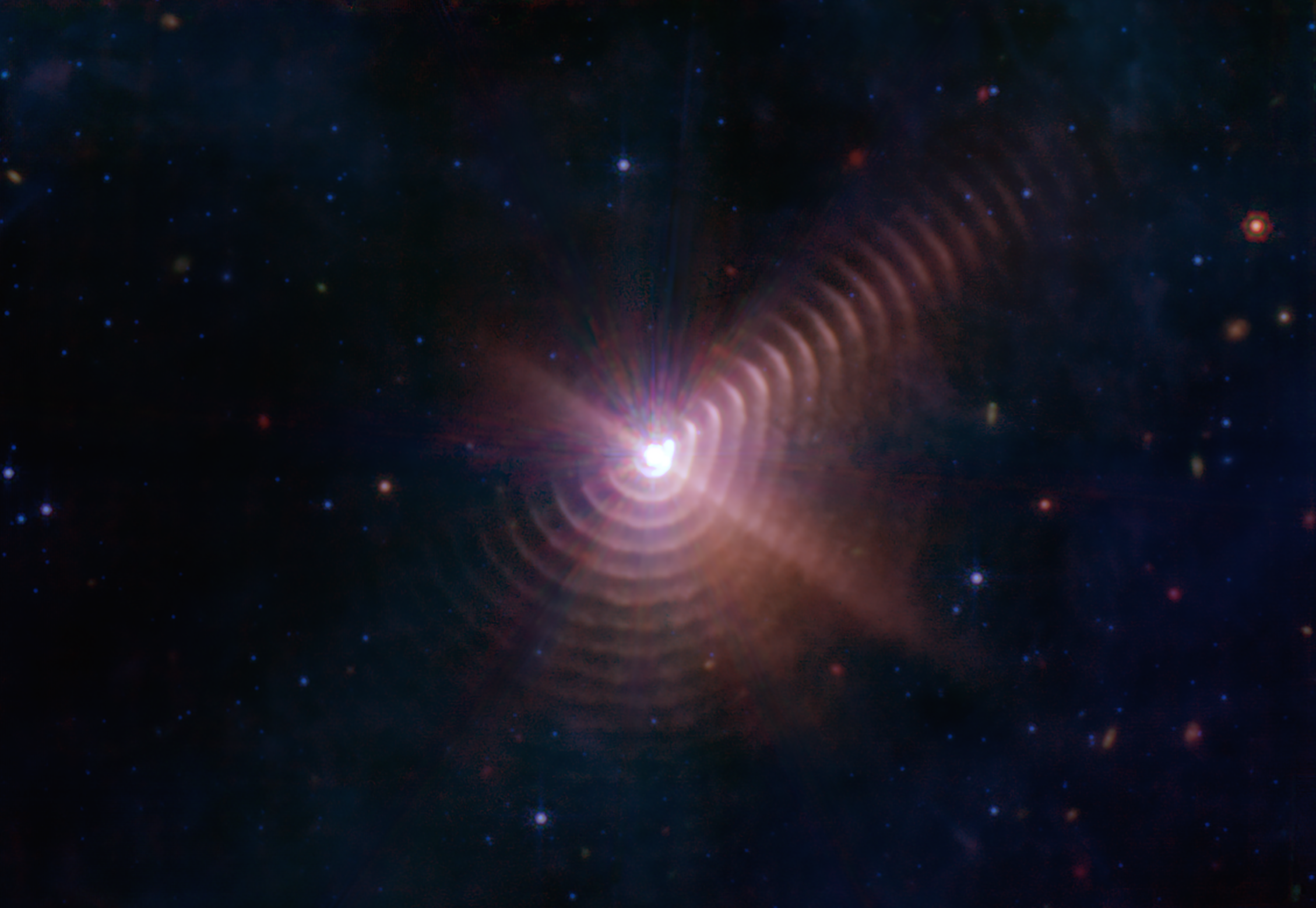 A bright white point of light is surrounded by ten to fifteen regularly spaced, hazy rings at its bottom, right, and upper right. The central point, where the stars are located, has a rough hexagon shape. The innermost ring is highlighted blueish white and is much brighter to the right. The outer rings fade from view to the upper left, with only a few close rings visible there. The central light seems to highlight the misshapen rings like a spotlight, with rays coming out diagonally from the upper left to lower right. One ray illuminates even more rings as it travels to the upper right.