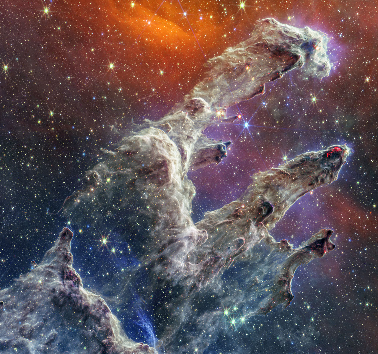 Layers of semi-opaque brown- and purple-colored gas and dust that starts at the bottom left and goes toward the top right. There are three prominent pillars rising toward the top right. The top pillar is the largest and widest. The peaks of the second and third pillars are set off in darker shades of brown and have red areas.