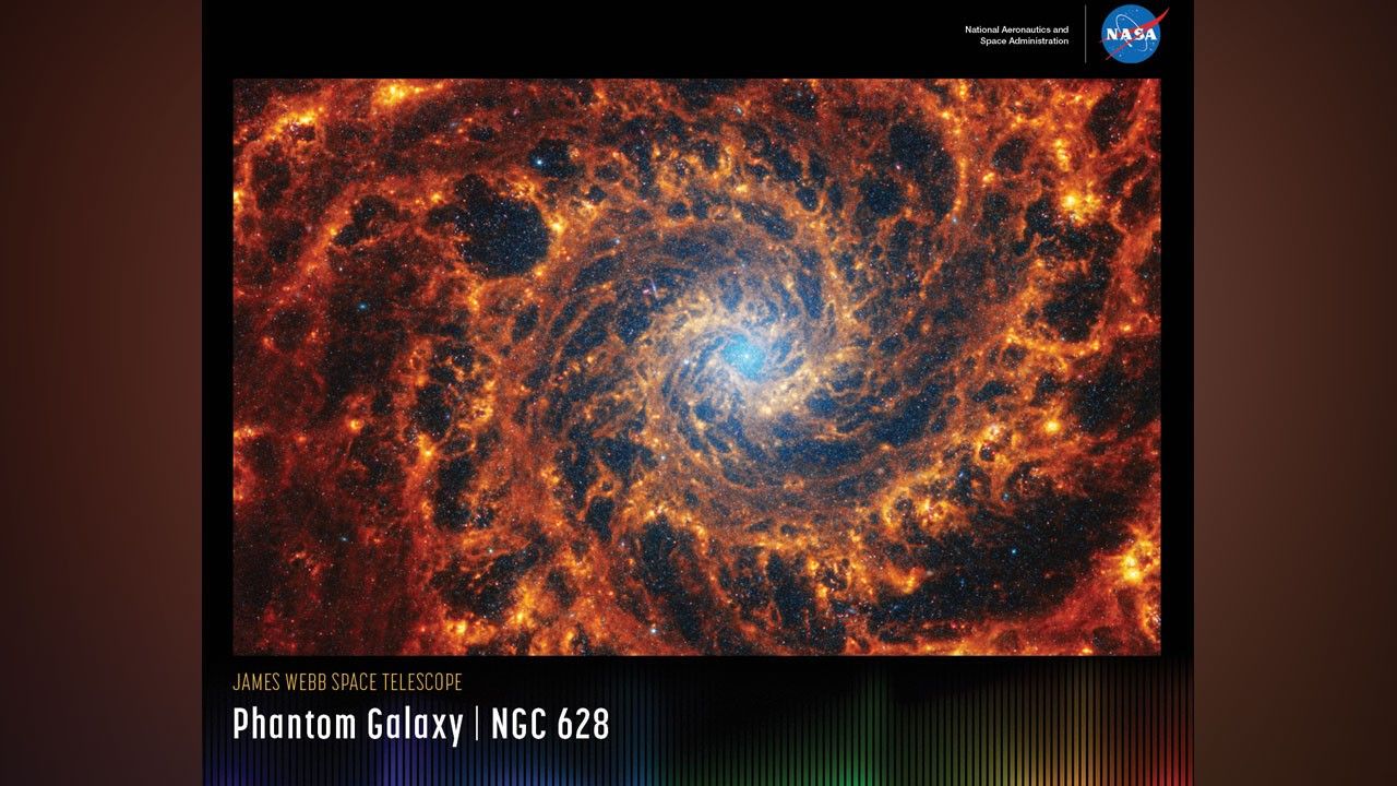 A horizontal poster displaying an infrared image of NGC 628. The galaxy's blue core, composed of older stars, is roughly centered. Its two spiral arms, which contain black pockets of different sizes, appear to rotate counterclockwise. The arms are composed of many filaments in different shades of orange, looking like a cross section of a nautilus shell. Brighter orange pockets indicate star formation. At upper right is text that reads, "National Aeronautics and Space Administration" and NASA's blue, white and red "meatball" logo. At lower left, the title text "James Webb Space Telescope" appears in small orange type on the top line with "Phantom Galaxy – NGC 628" in large white type below. Along the bottom is a series of thin vertical bars ranging from purple at left to blue, green, yellow, orange, and red at right. Bars are separated slightly, with black in between. Bar colors are brightest at the bottom, fading up to black. Brightness varies from bar to bar, giving the array an undulating feel.