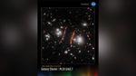 A vertical poster with an image of PLCK G165.7 against a black background. At left of center of the image, there is a red arc that stretches from 2 to 7 o’clock. At its left is a cluster of a few white galaxies. At right of the center, there are red arcs stretching from 3 to 5 o’clock, and a set of white galaxies at their right. There are white, blue, and red galaxies spread throughout with a few arc-shaped galaxies near the white galaxy clusters. Along the bottom is a series of thin vertical bars ranging from purple at left to blue, green, yellow, orange, and red at right. Bars are separated slightly, with black in between. Bar colors are brightest at the bottom, fading up to black. Brightness varies from bar to bar, giving the array an undulating feel. At upper right is text that reads, "National Aeronautics and Space Administration" and NASA's blue, white, and red meatball insignia. Title text appears at lower left that says, "James Webb Space Telescope: Galaxy Cluster, PLCK G165.7."