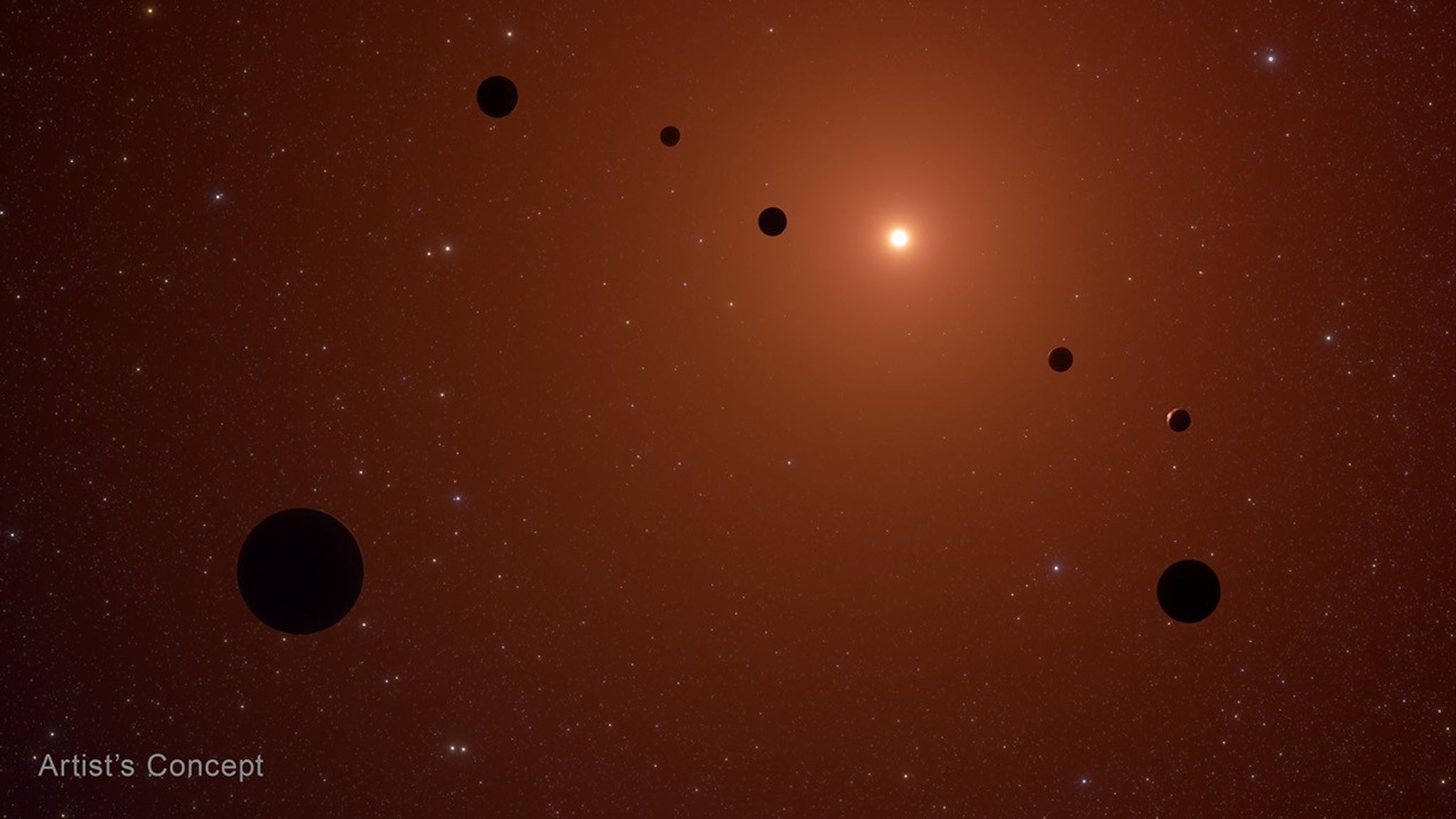 Artist’s concept of the TRAPPIST-1 system, which is known to have seven Earth-sized rocky planets orbiting a red dwarf star. The host star, located in the background, is depicted as a bright white circle. It is surrounded by a yellow glow that fades to orange near the edges of the frame. The seven planets appear in the foreground, with their nightsides, the side facing away from the host star, facing the viewer. The planets are depicted as red-brown spheres. Many distant galaxies and stars faintly appear in the background. Text in bottom left corner reads “Artist’s Concept.”