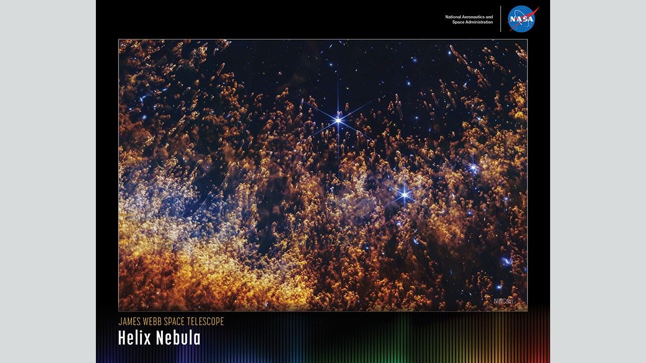 A horizontally oriented poster with a portion of the Helix Nebula against a black background. Thousands of orange and gold comet-like pillars stream upward from the bottom, which are organized in an orange, semi-circular arc. The pillars are denser and orange at the bottom, but thinner and more golden toward the top. The background of space is more visible toward the top. A few large blue stars are on the right side. Along the bottom of the poster is a series of thin vertical bars ranging from purple at left to blue, green, yellow, orange, and red at right. These bars are separated slightly, with black in between. Bar colors are brightest at the bottom, fading up to black. Brightness varies from bar to bar, giving the array an undulating feel. At upper right is text that reads, "National Aeronautics and Space Administration” and NASA’s blue, white, and red meatball insignia. Title text appears at lower left that says, “James Webb Space Telescope: Helix Nebula."