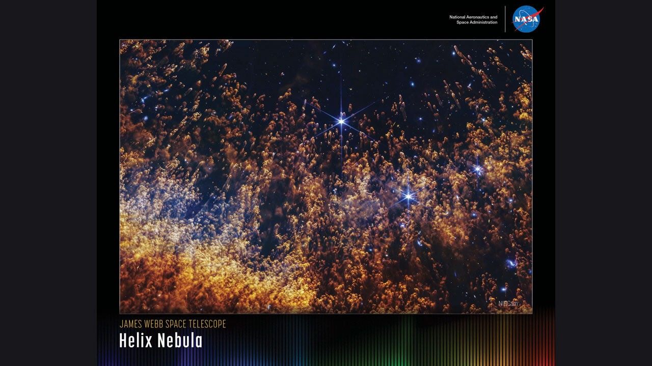 A horizontally oriented poster with a portion of the Helix Nebula against a black background. Thousands of orange and gold comet-like pillars stream upward from the bottom, which are organized in an orange, semi-circular arc. The pillars are denser and orange at the bottom, but thinner and more golden toward the top. The background of space is more visible toward the top. A few large blue stars are on the right side. Along the bottom of the poster is a series of thin vertical bars ranging from purple at left to blue, green, yellow, orange, and red at right. These bars are separated slightly, with black in between. Bar colors are brightest at the bottom, fading up to black. Brightness varies from bar to bar, giving the array an undulating feel. At upper right is text that reads, "National Aeronautics and Space Administration" and NASA’s blue, white, and red meatball insignia. "Title text appears at lower left that says, "James Webb Space Telescope: Helix Nebula."