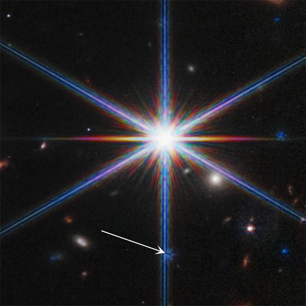 A bright star with eight thicker, blue-white diffraction spikes appears almost at center. A few stars and galaxies are in the background, which is black. A white arrow points to another tiny star within the large star’s bottom-center diffraction spike. That tiny star also has its own diffraction spikes, but is an artifact from a previous exposure.