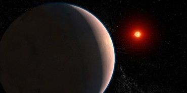 Occupying the left two-thirds of the image is a foreground planet mostly in shadow. On the right side, a tan crescent shows subtle surface features. A thin, tenuous blue atmosphere lines the planet’s limb. On the right, a small red globe represents a red dwarf star. Its surface is mottled with small, dark spots resembling sunspots. Both planet and star are on a mostly black background speckled with hundreds of faint, distant stars.