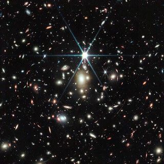 A black background is scattered with hundreds of small galaxies of different shapes, ranging in color from white to yellow to red. Some galaxies, mostly the redder galaxies, are distorted, appearing to be stretched out or mirror imaged. Just a little bit above the center, there is a bright source of light, a star, with 8 bright diffraction spikes extending out from it. Below the star are several noticeably fuzzy white galaxies that resemble cotton balls – these are part of a galaxy cluster. To the lower right of the star is a particularly long, red, thin line stretching from one o’clock to 7 o’clock. This is the Sunrise Arc.