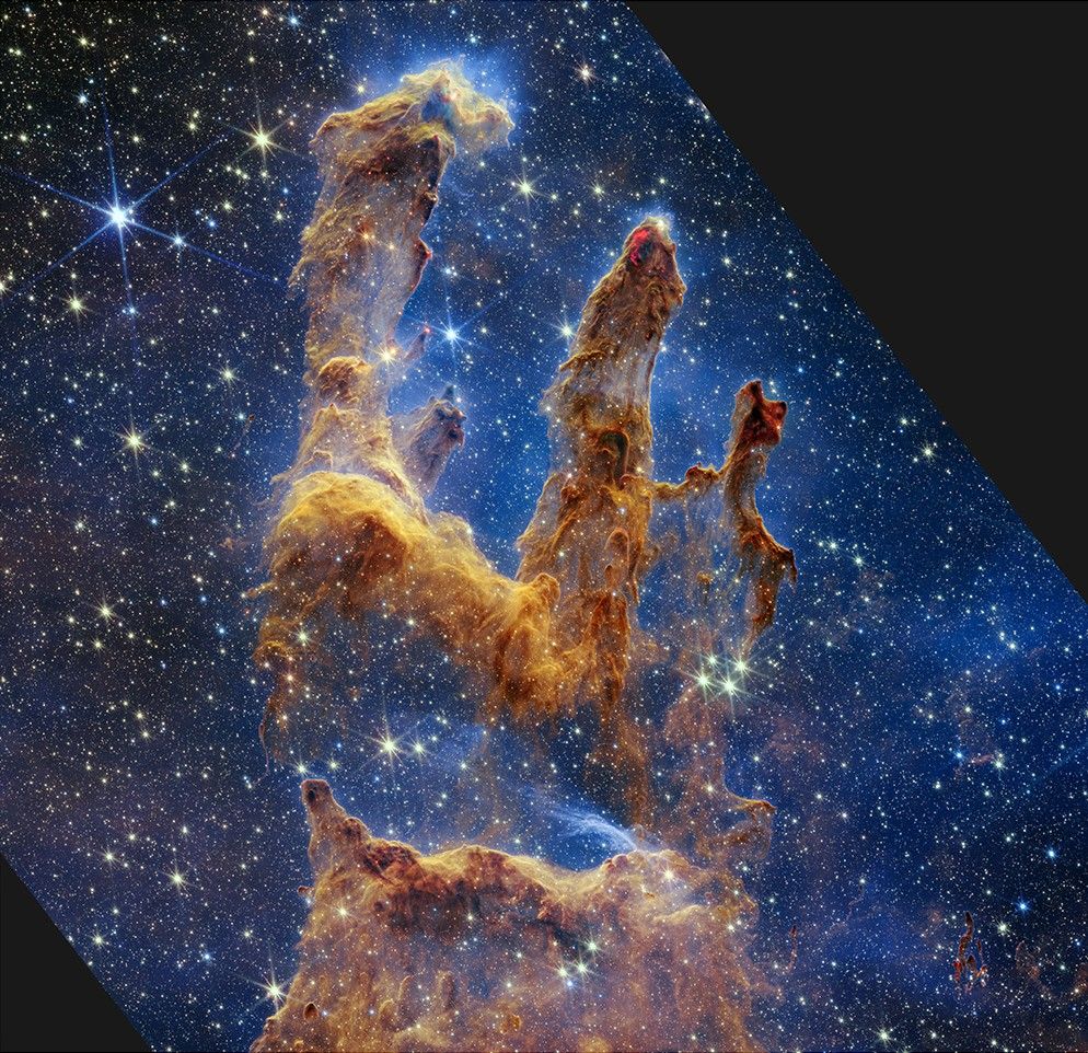 At the bottom left of this vertical image are the thickest regions of brown and rusty red gas and dust. There are many layers of semi-transparent gas and dust overlaying one another. A peak rises about a third of the way from the bottom, and becomes far darker brown with two bright red areas toward the tip. The light brown dust becomes more diaphanous about halfway up the screen. There’s a slight gap in the dust, which allows the blue background to come into view clearly. About 60% of the background in this image is set in shades of blue and littered with tiny yellow and blue stars. The brown pillars continue, taking the shape of a shoulder at the base, with three prominent columns rising out toward the upper right. The top left pillar is the largest and widest. The peaks of the second and third pillars are set off in darker shades of brown and have red outlines. For more details, download the Text Description.