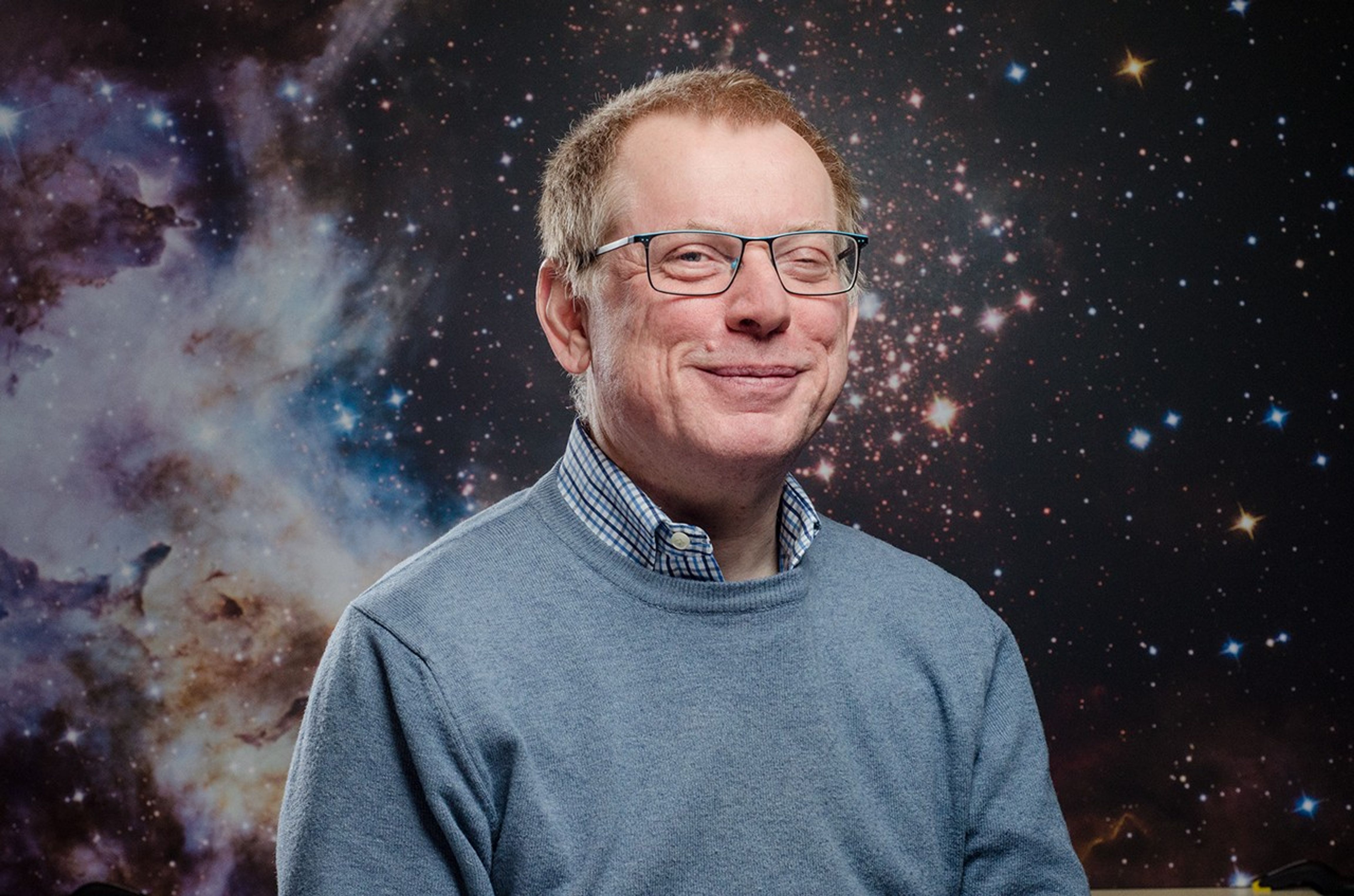 White man smiles in front of a cosmic background. He has light brown short hair and has black and blue-colored glasses. He is wearing a checkered collared shirt and a light blue sweater.