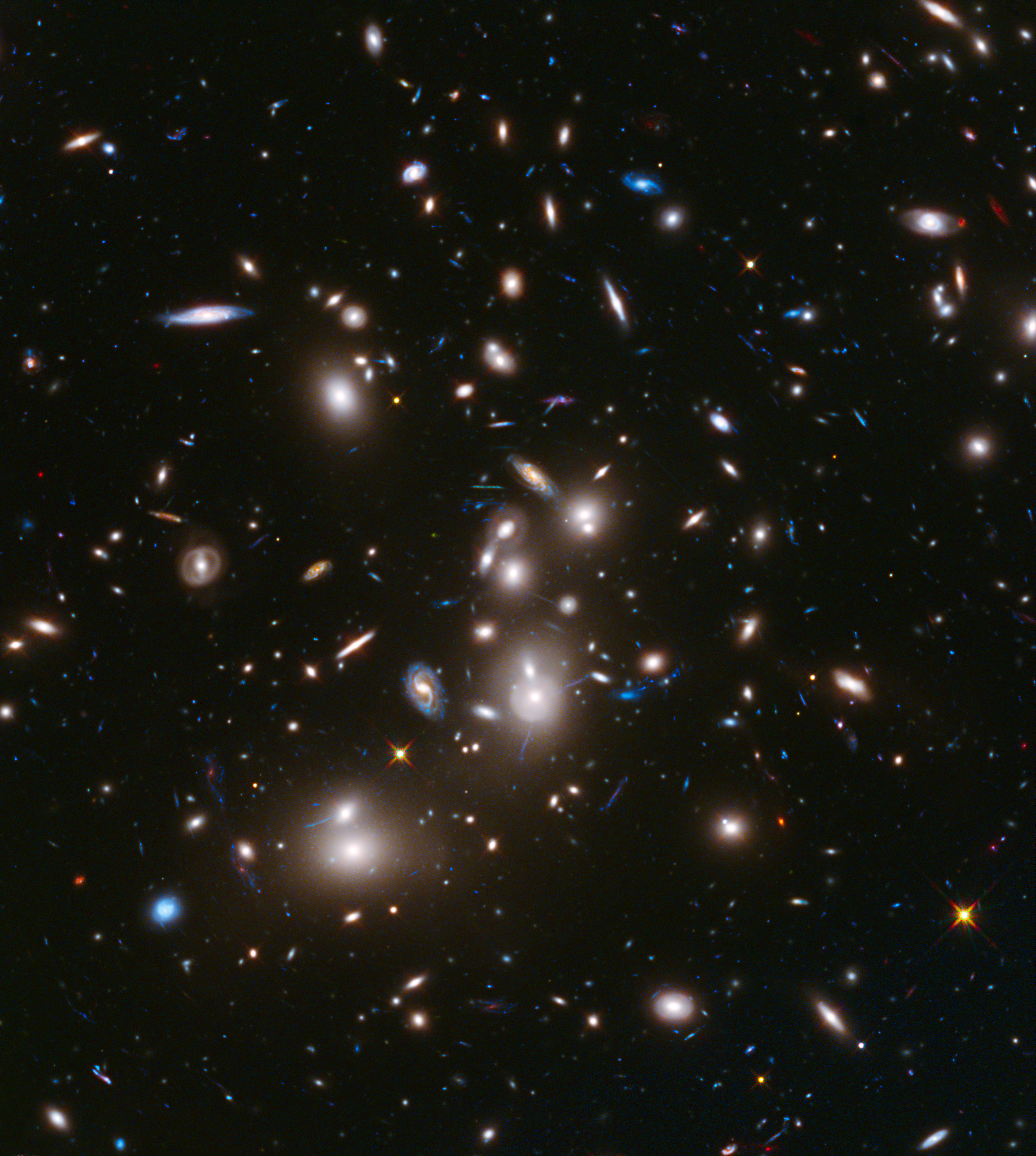Galaxy cluster Abell 2744. Many yellow and white galaxies appear across this black swath of space, with the largest appearing toward the center. Small, thin blue-white lines and tiny red points of light are peppered across the image.