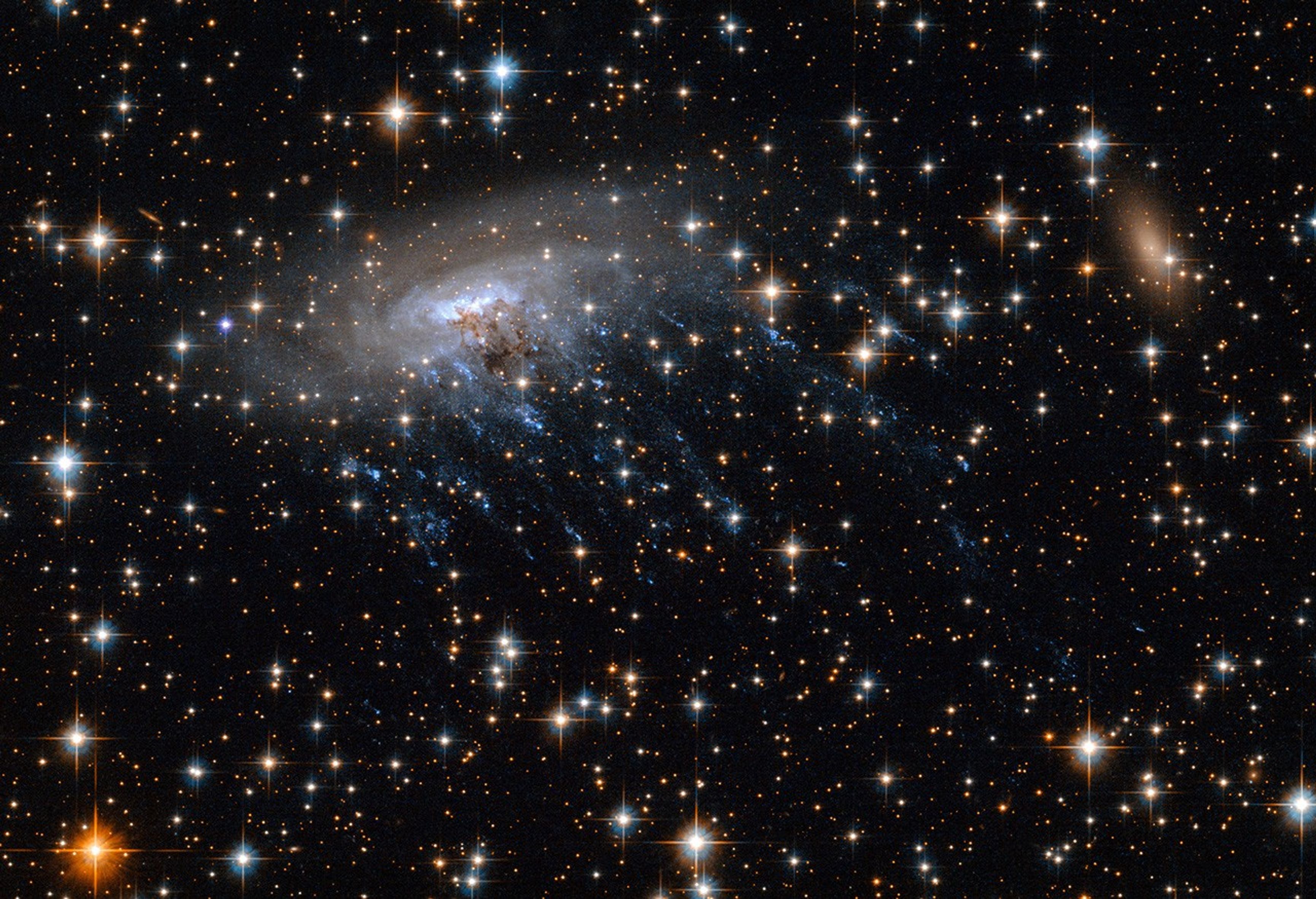 Left of center is a galaxy that looks like a white-and-blue swirl with several blue filaments that trail downward. Its transparent white spiral arms rotate in a clockwise motion.