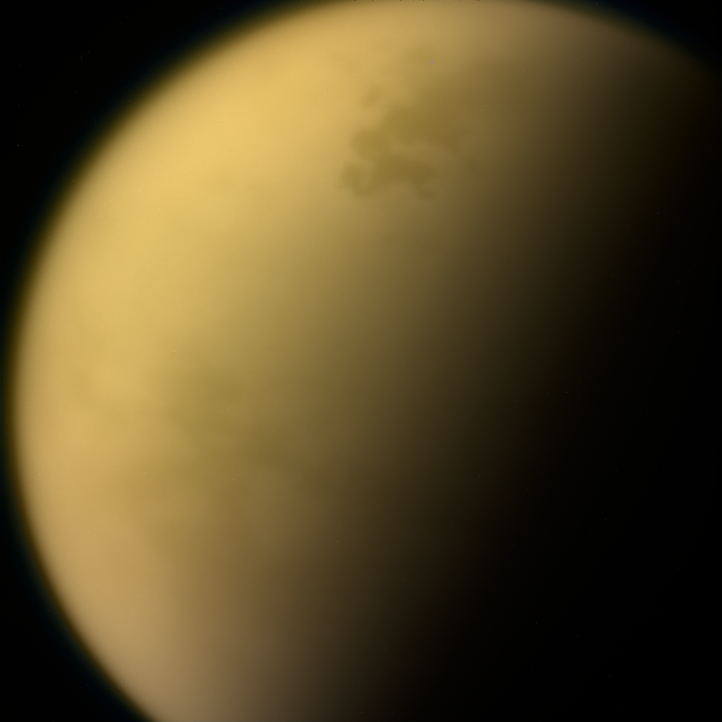 A close-up of Saturn's moon Titan as imaged by the Cassini spacecraft. Against a black background is a large, fuzzy sphere. Its north and south regions extend beyond the frame. The left side of the moon is roughly illuminated, revealing a slightly uneven yellow surface. The right side of the moon is shrouded in darkness.