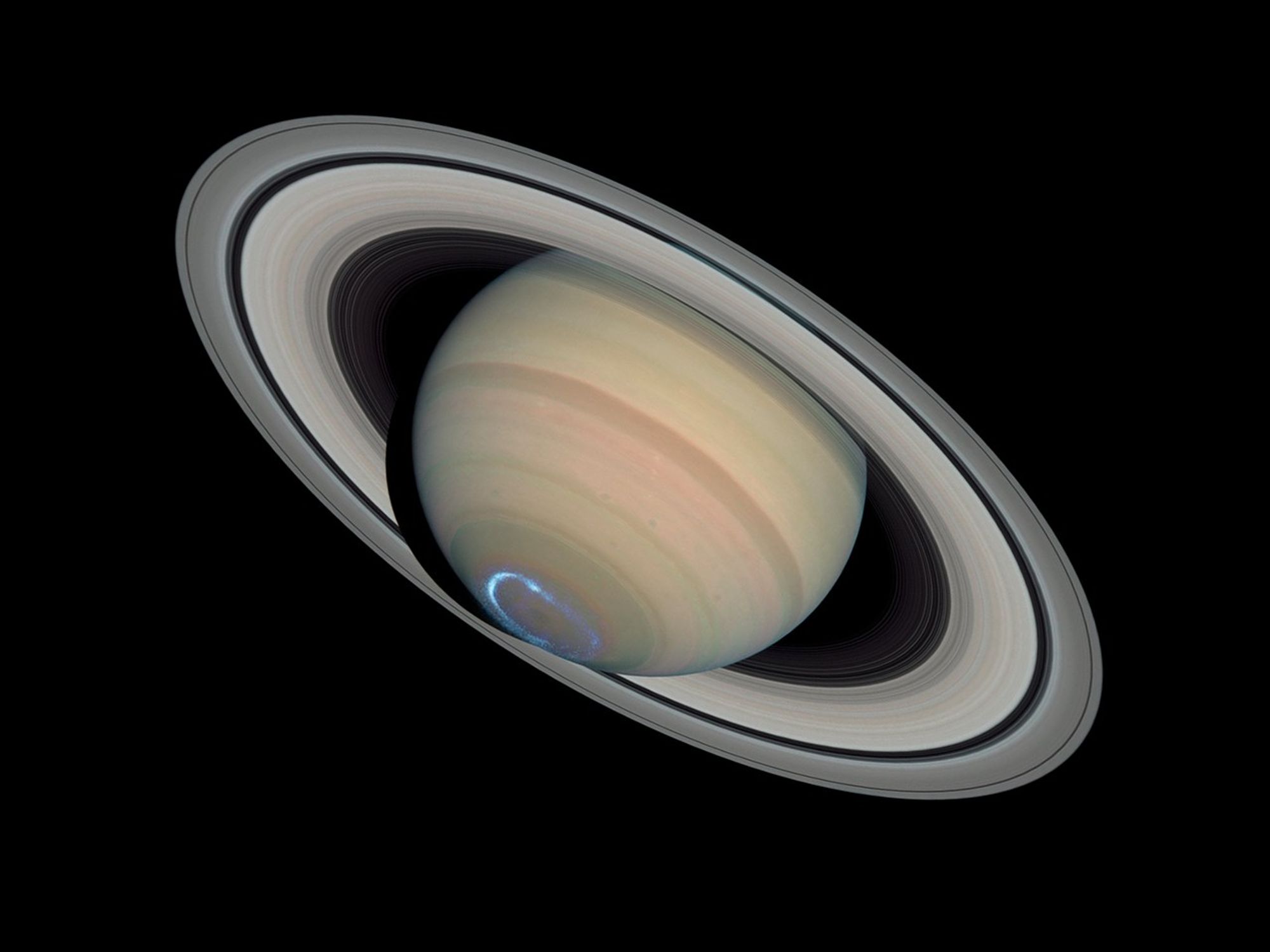 Saturn against the black background of space. The planet is made up of a very large sphere at the center, which has a range of beige, muted pink, and light green bands around it. It is surrounded by a system of rings, which extends from the top left to the bottom right of this image.