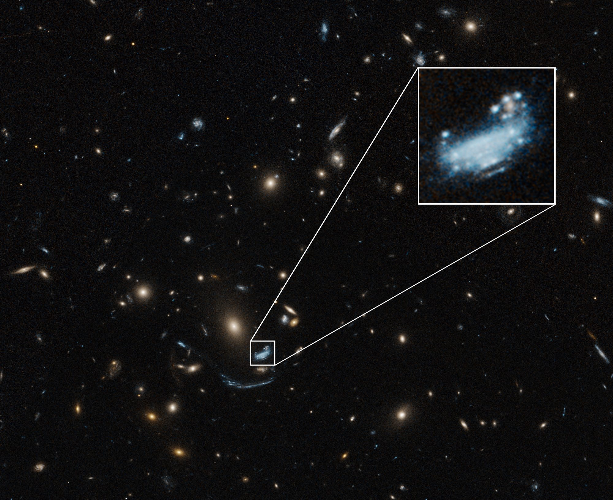 A largely black image is covered with blue, white, yellow, and orange dots of various sizes throughout. Toward the bottom center, a blue galaxy is highlighted by a small white square, which is enlarged near the top right corner.