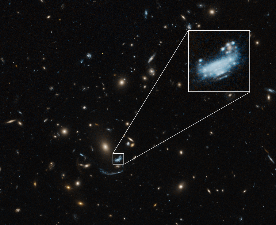 A largely black image is covered with blue, white, yellow, and orange dots of various sizes throughout. Toward the bottom center, a blue galaxy is highlighted by a small white square, which is enlarged near the top right corner.
