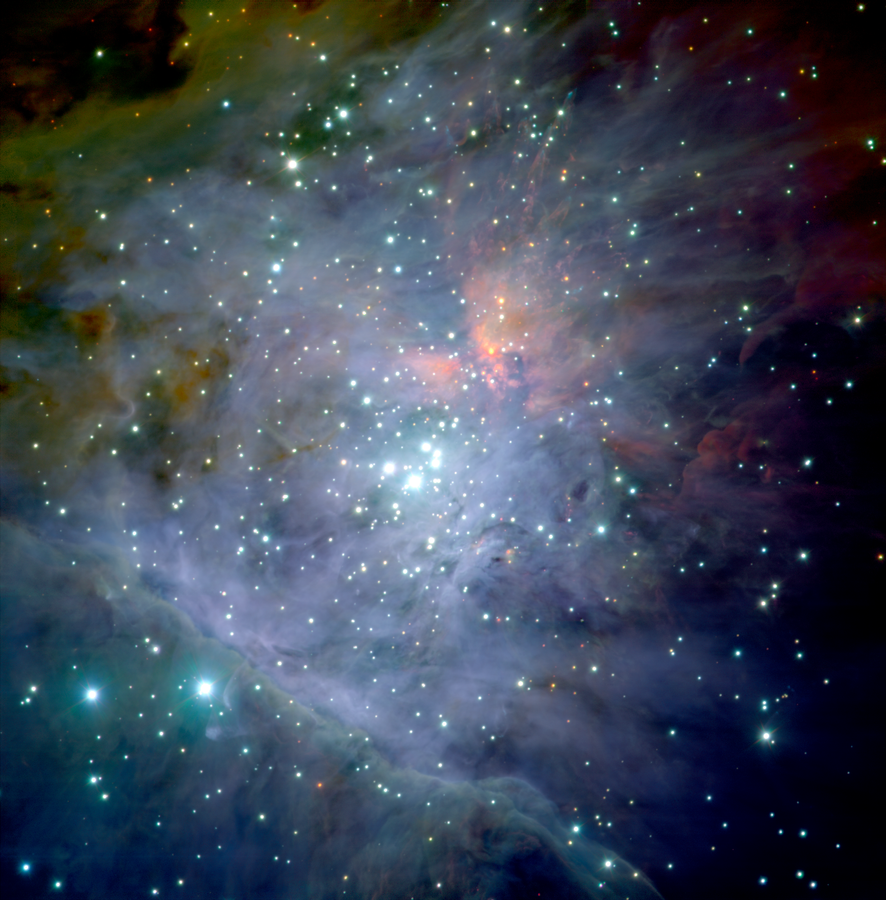 About a thousand young stars, which are mostly light blue, are crammed into the scene. The bulk of the image is awash in light purple, which takes irregular shapes and appears wispy, but there’s a pink splotch at top center, and a yellow-green region at top left.