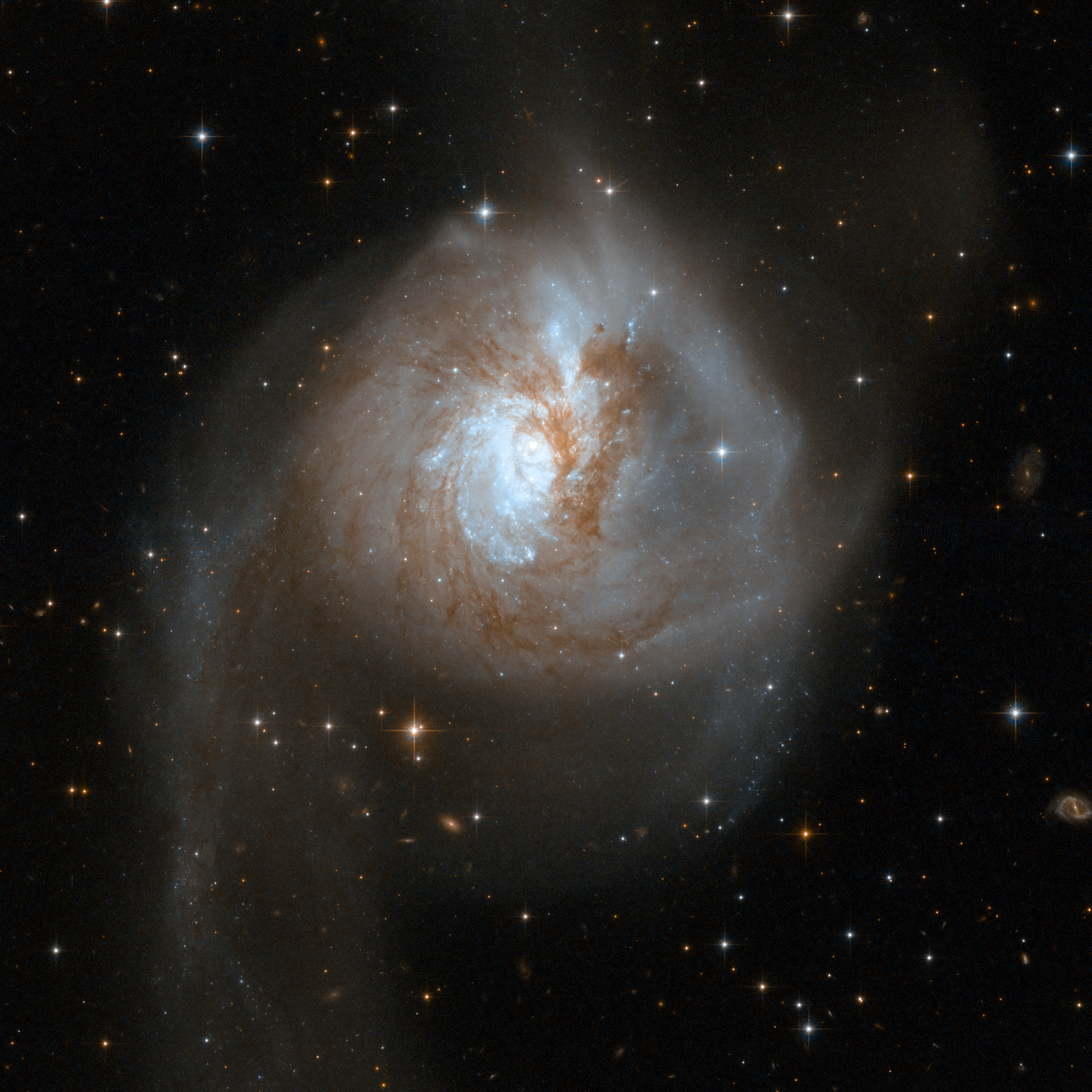 NGC 3256, a pair of interacting galaxies with a large egg-like shape. Brown-red dust is mixed throughout the center, and two uneven light-blue “arms” extend down from the middle.